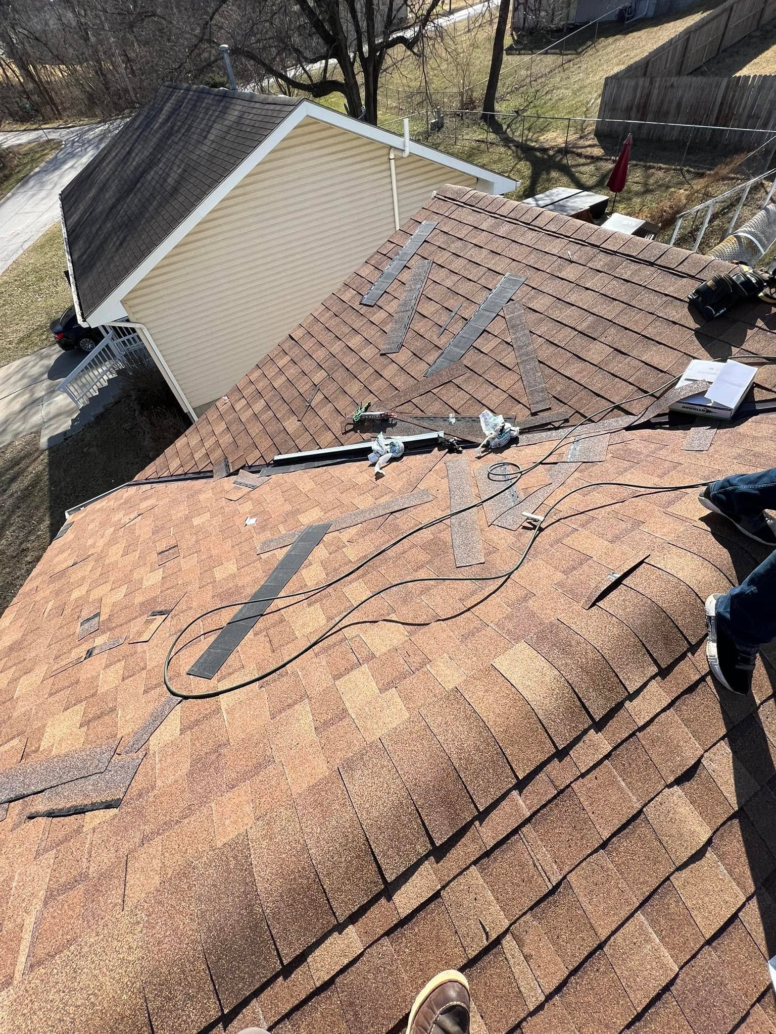 Damaged shingle roof with exposed underlayment and debris near a house