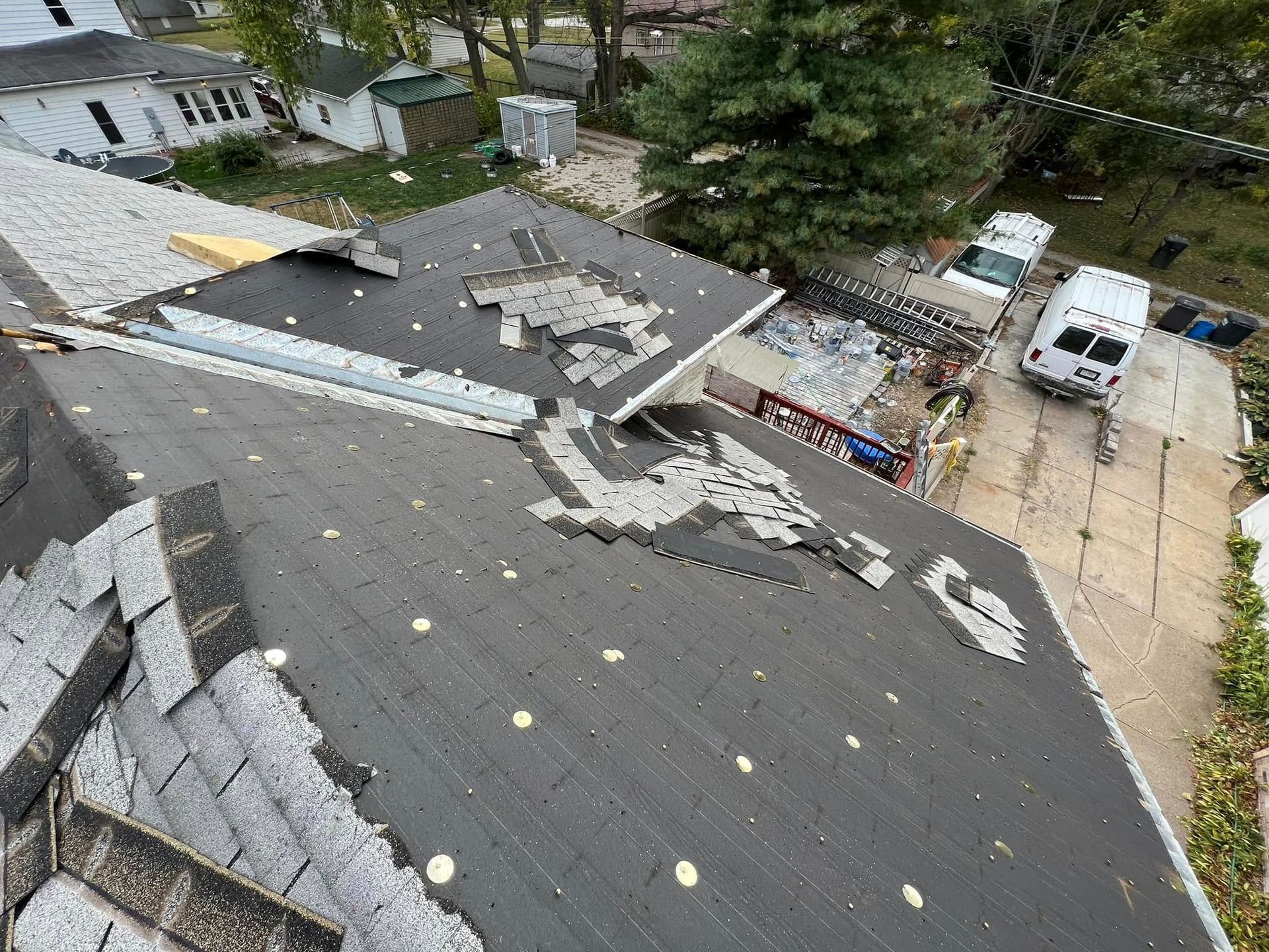 Damaged roof with scattered shingles and debris beside a driveway and white van in a wooded area