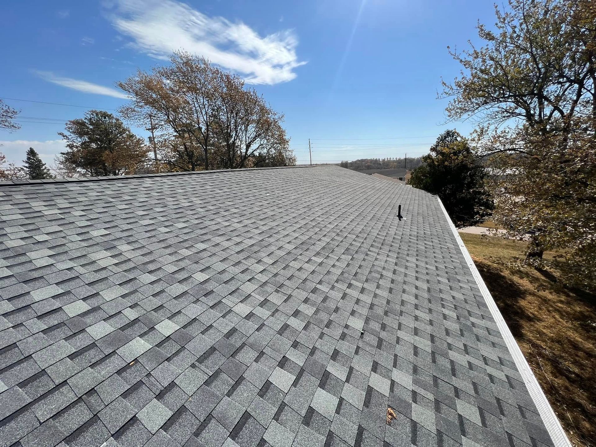 Gray shingled roof with a small vent under a blue sky and bare trees nearby