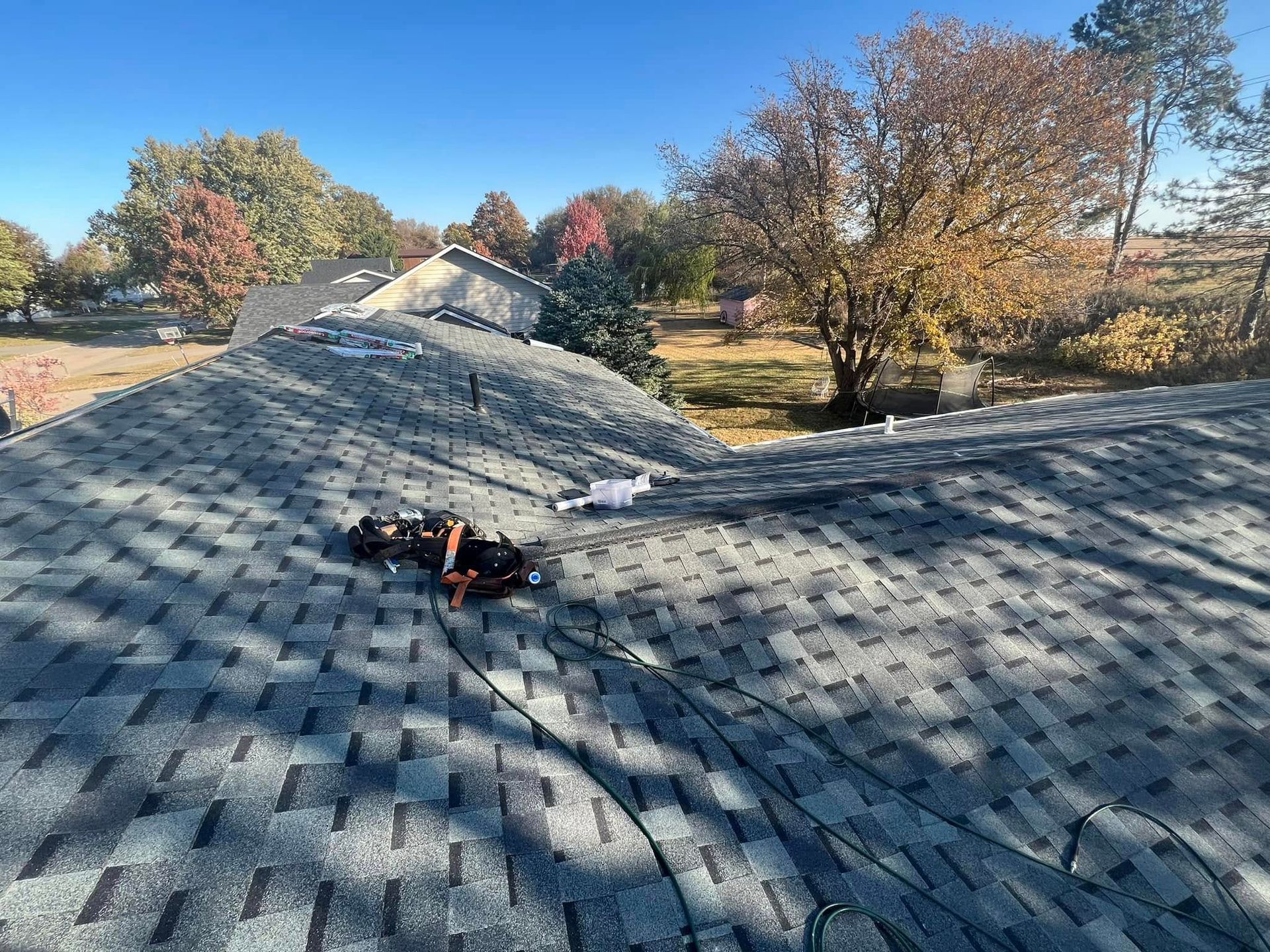Rooftop covered in gray shingles with a person lying near the ridge under clear autumn trees.