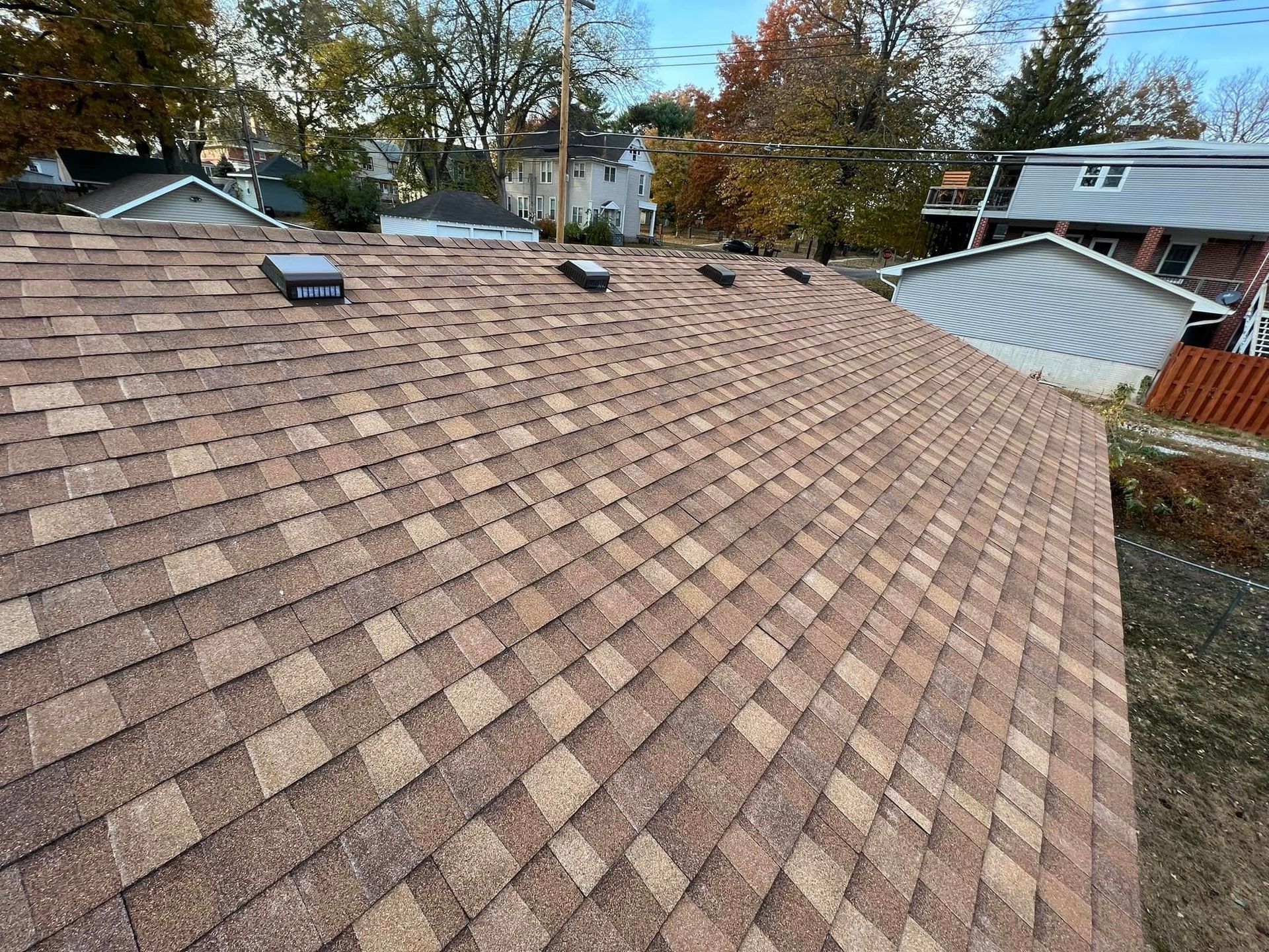 Brown shingled roof with a small vent, viewed from above with houses and autumn trees in the background