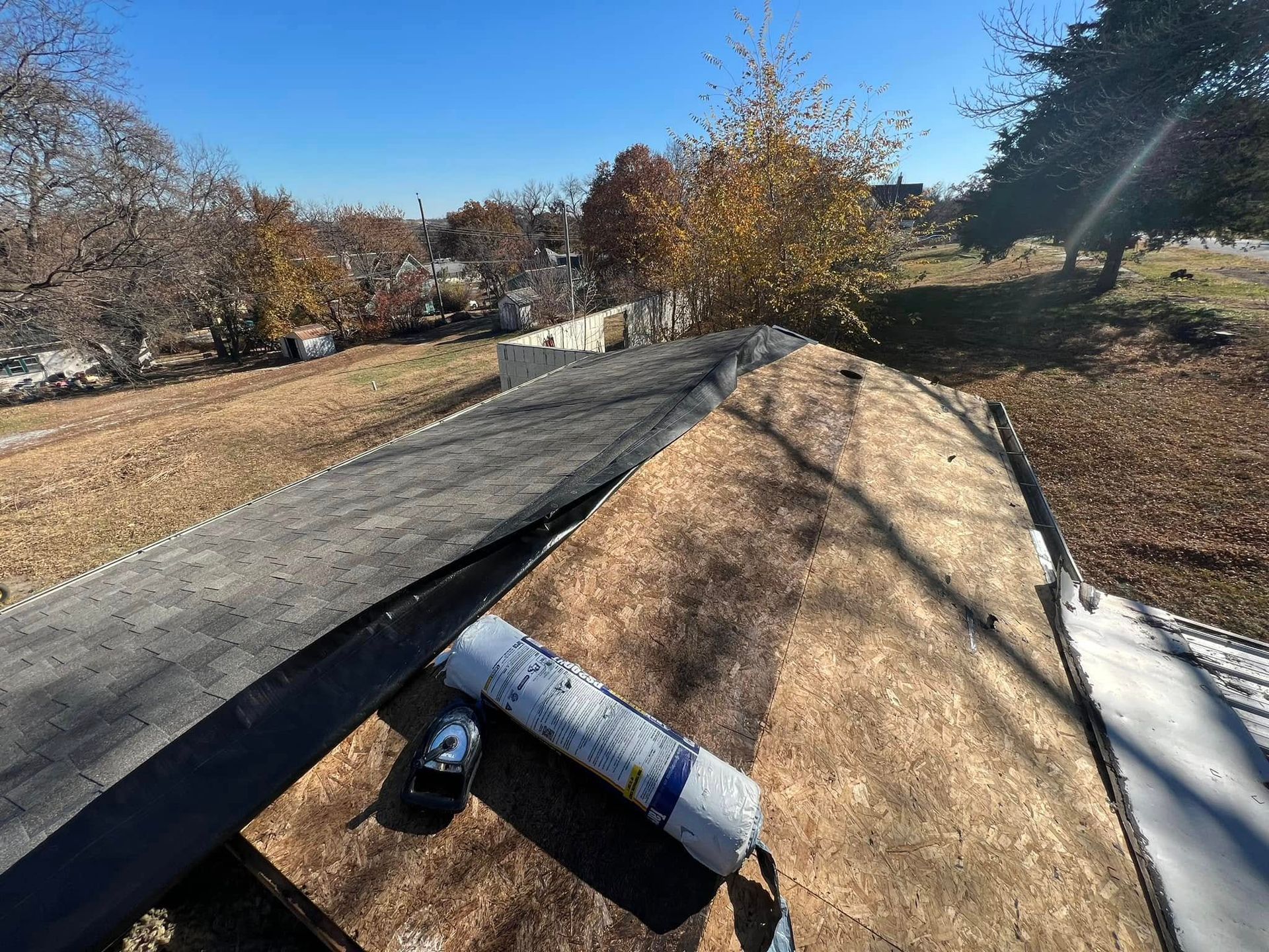 Rooftop under construction with plywood sheets, roofing material, and a wooded yard in the background