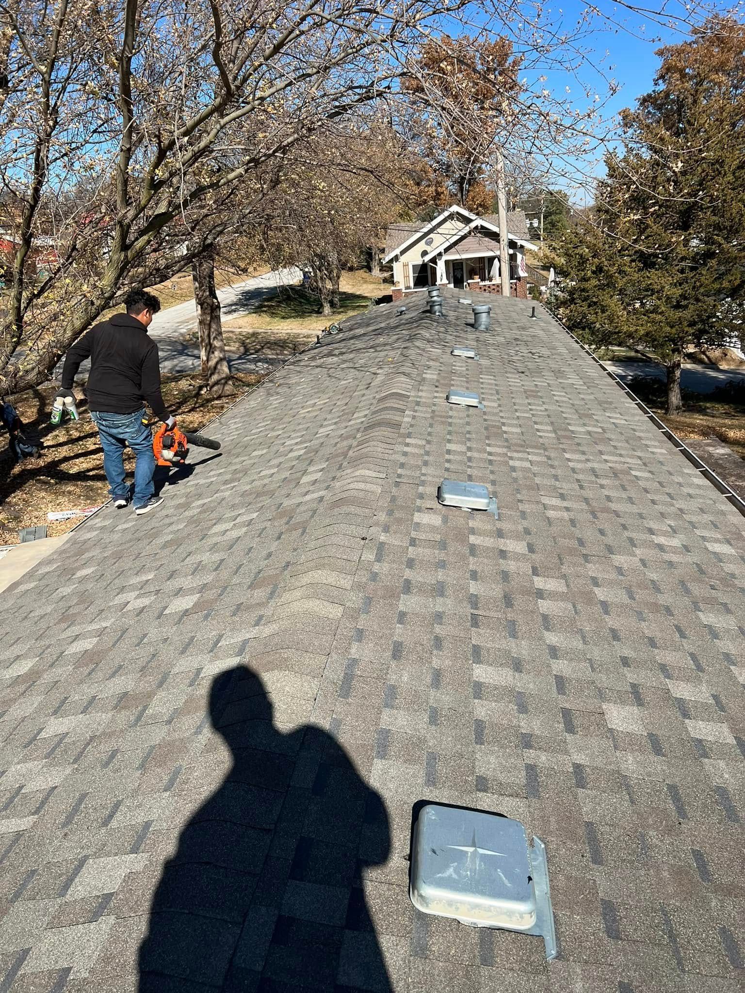 Worker walking on a shingled roof with vents, trees and a house in the background