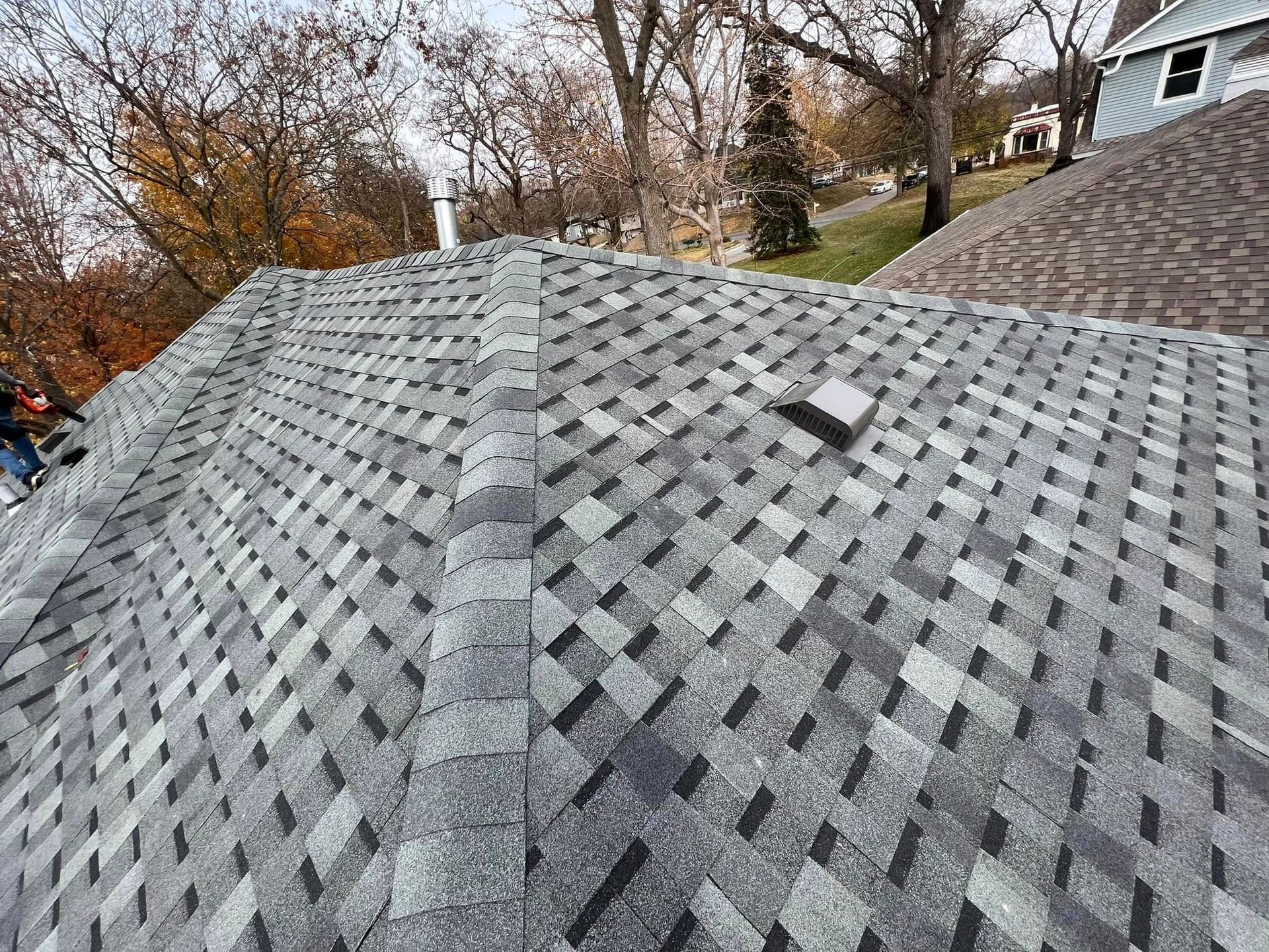 Gray shingled roof with a damaged patch and a roof vent, viewed from above among autumn trees