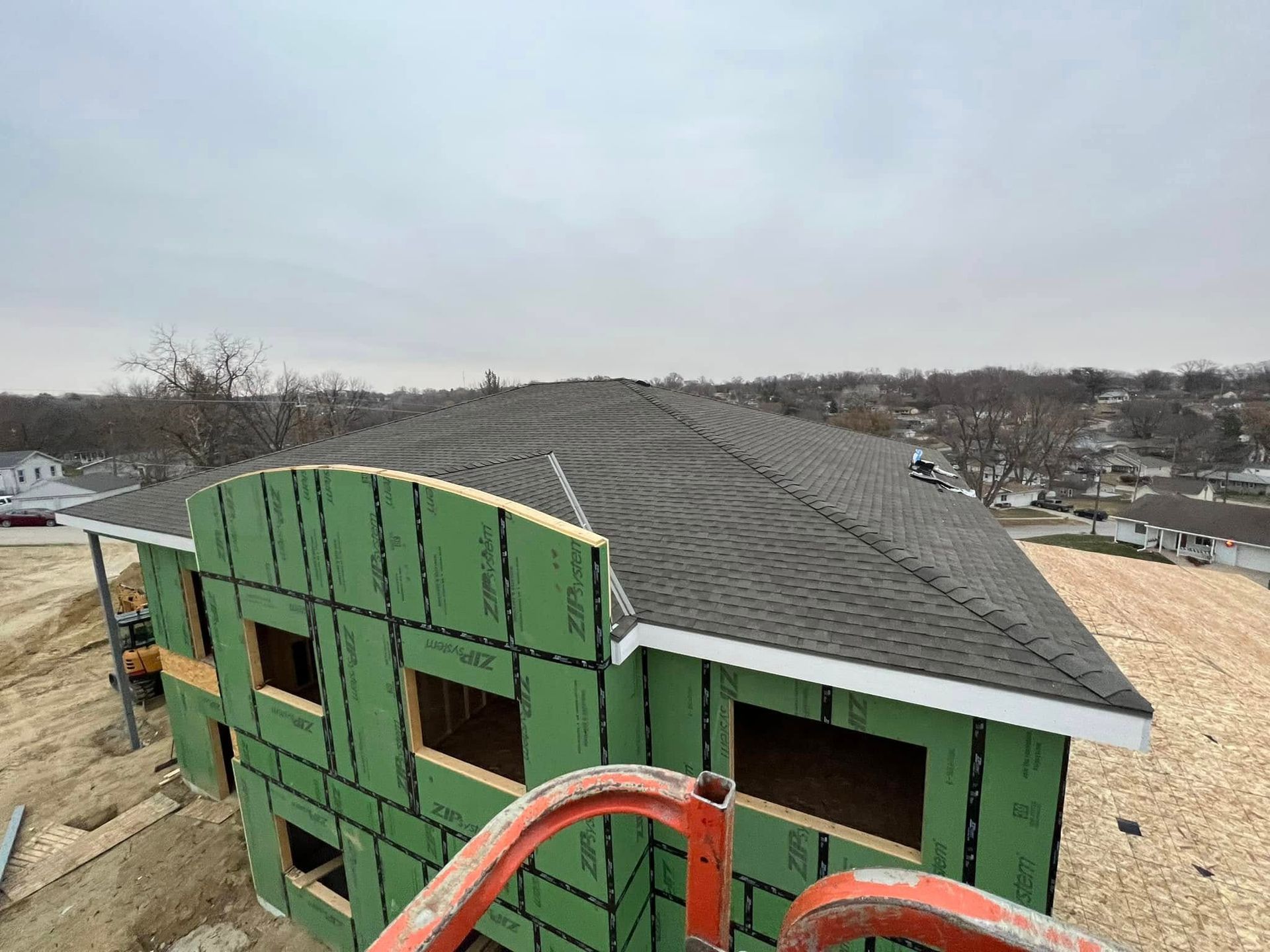 Partially built green house with workers and a red boom lift on a rooftop in a suburban area