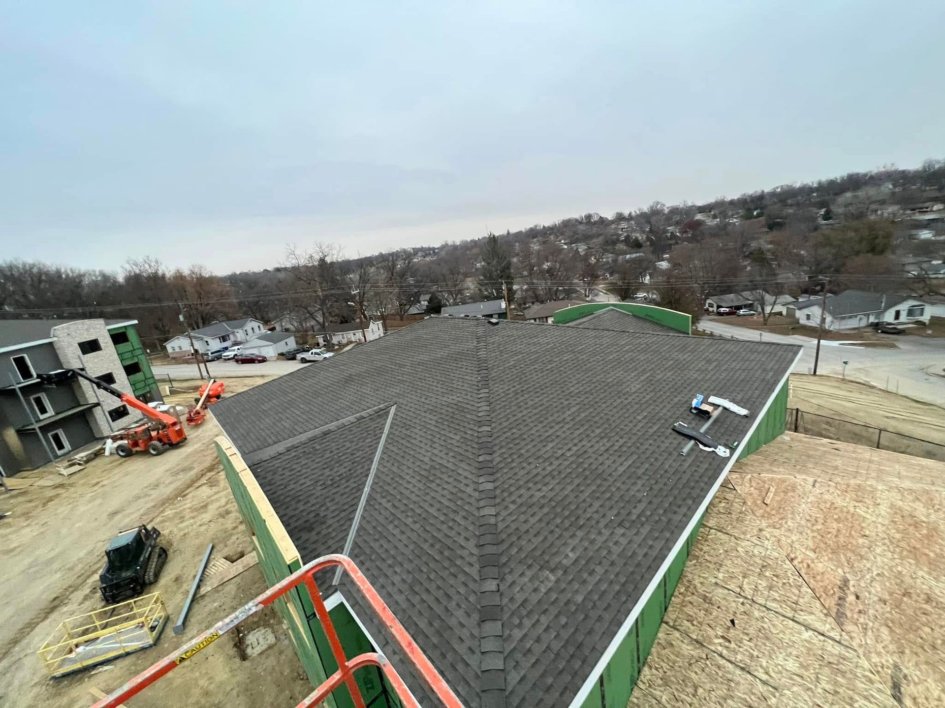 Rooftop under construction with gray shingles, surrounded by unfinished homes and dirt lots.