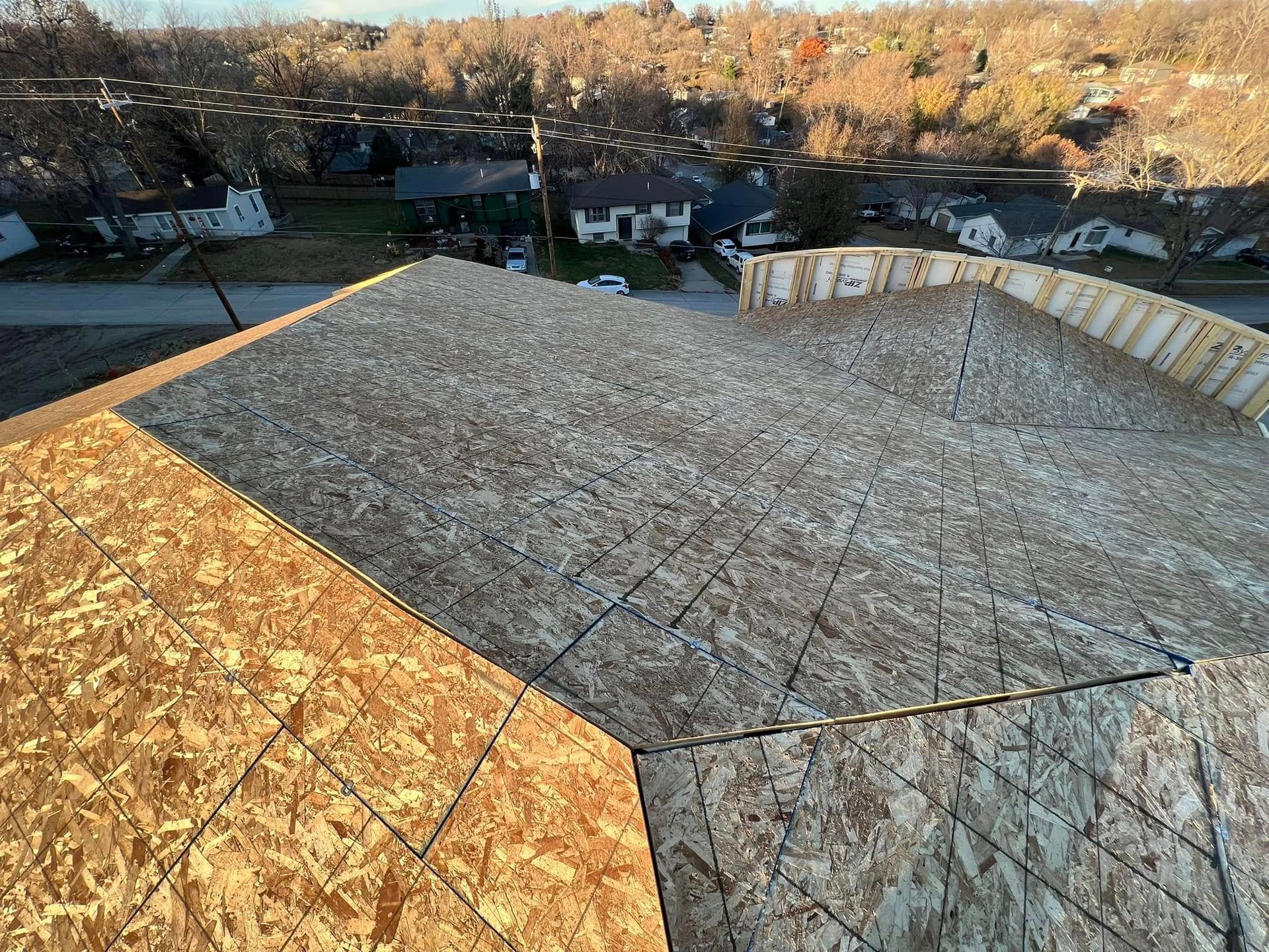 Partially built roof deck covered in plywood sheets, overlooking houses and trees at sunset