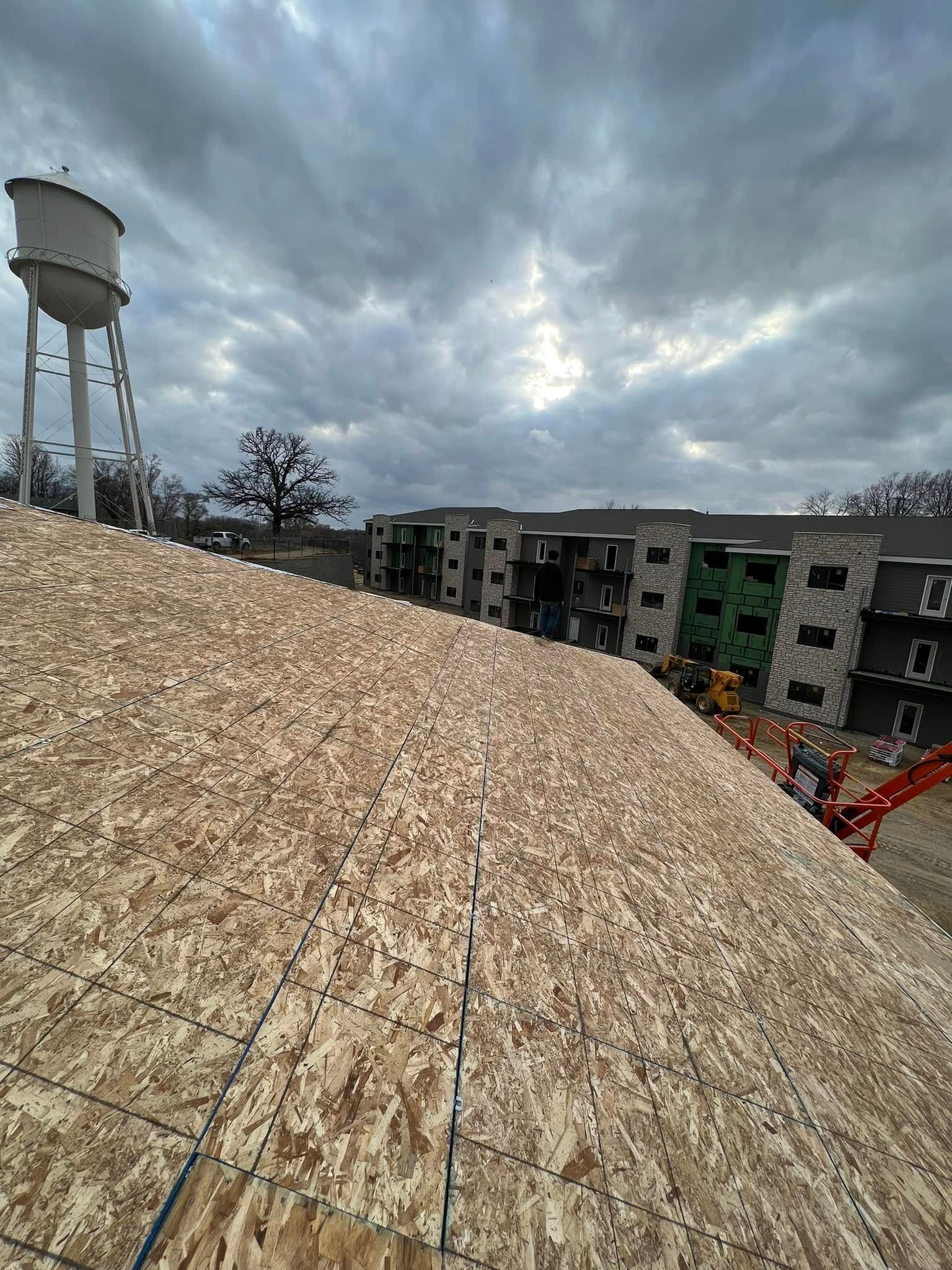 Rooftop under construction with plywood sheathing, a water tower, and cloudy sky over nearby buildings