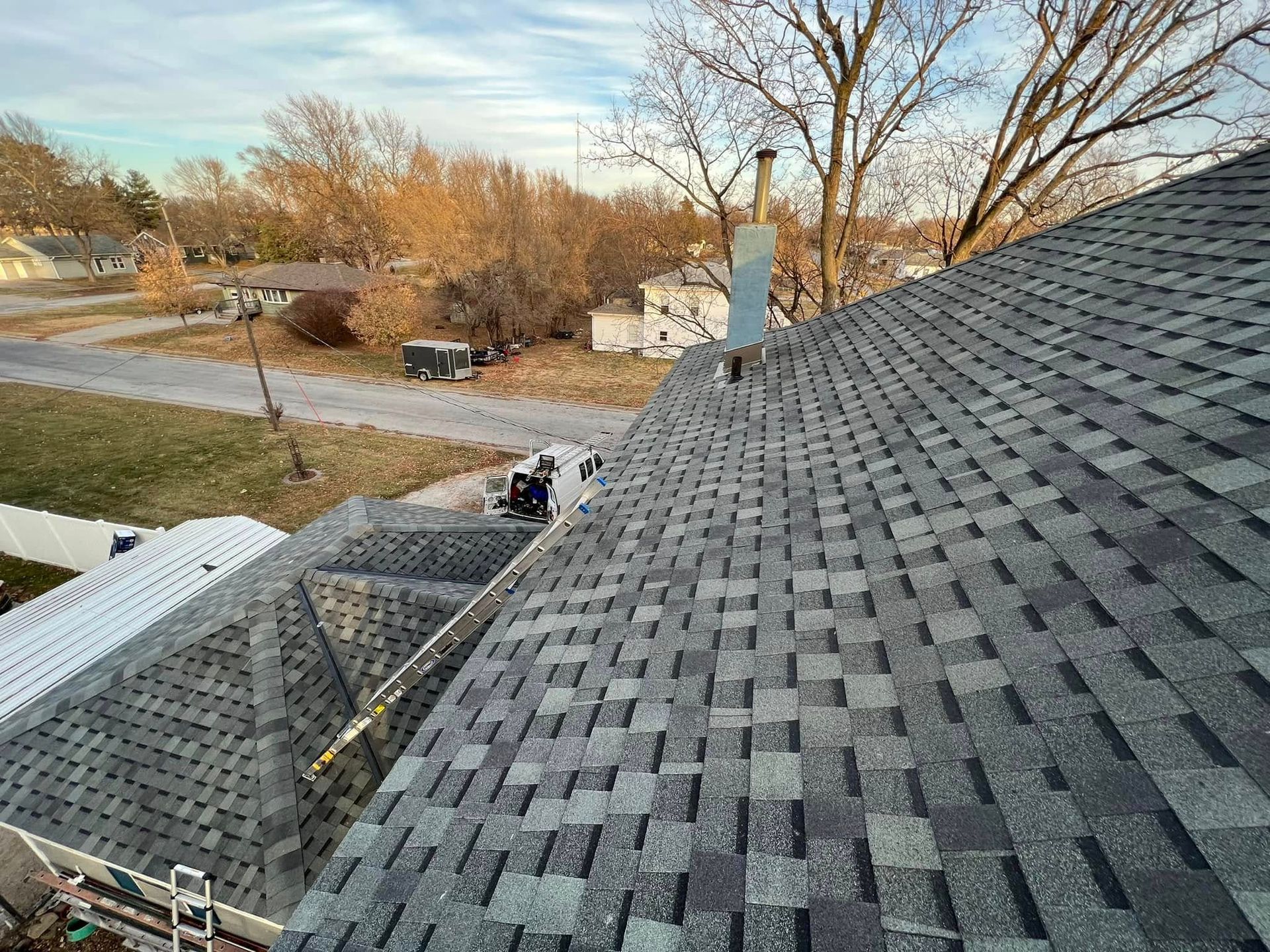 Roof shingles under repair with a worker on a sloped rooftop overlooking a quiet suburban street.