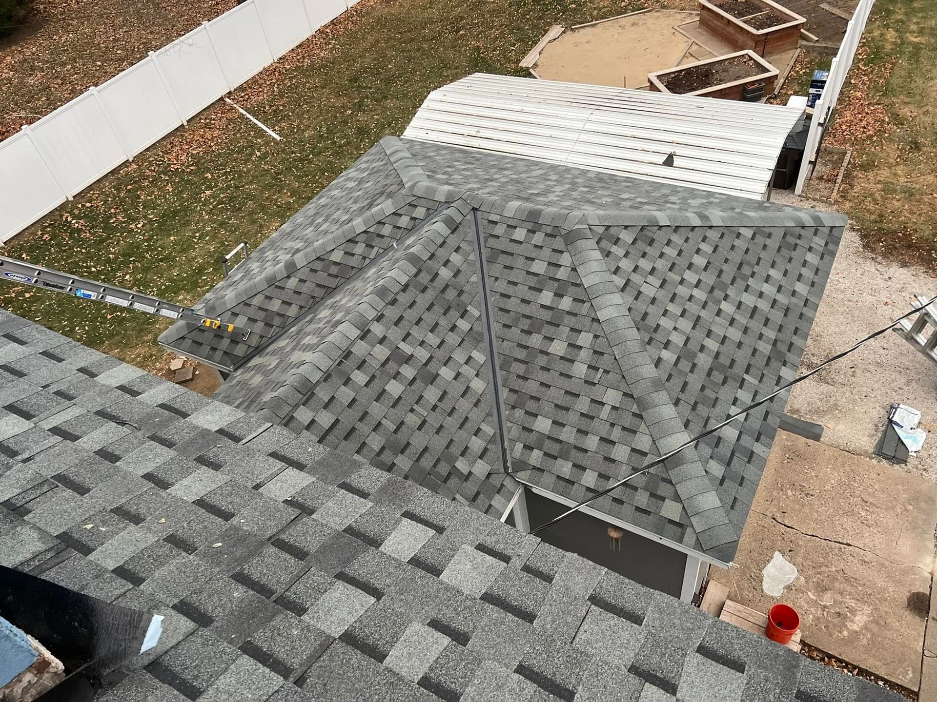 Roof under construction with gray shingles, viewed from above near a yard and wall.