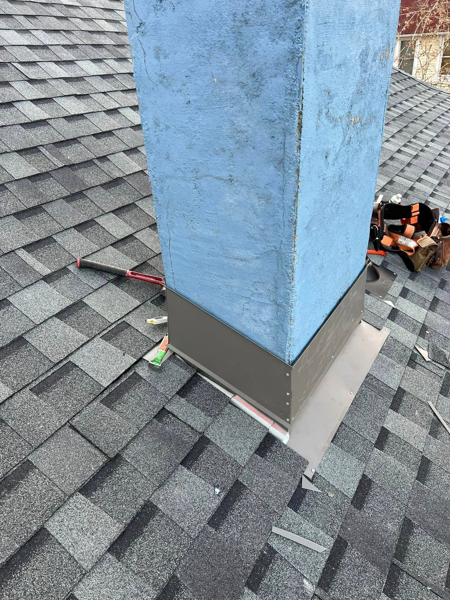 Blue chimney with gray flashing on a shingled roof, showing a roof repair area with exposed materials.