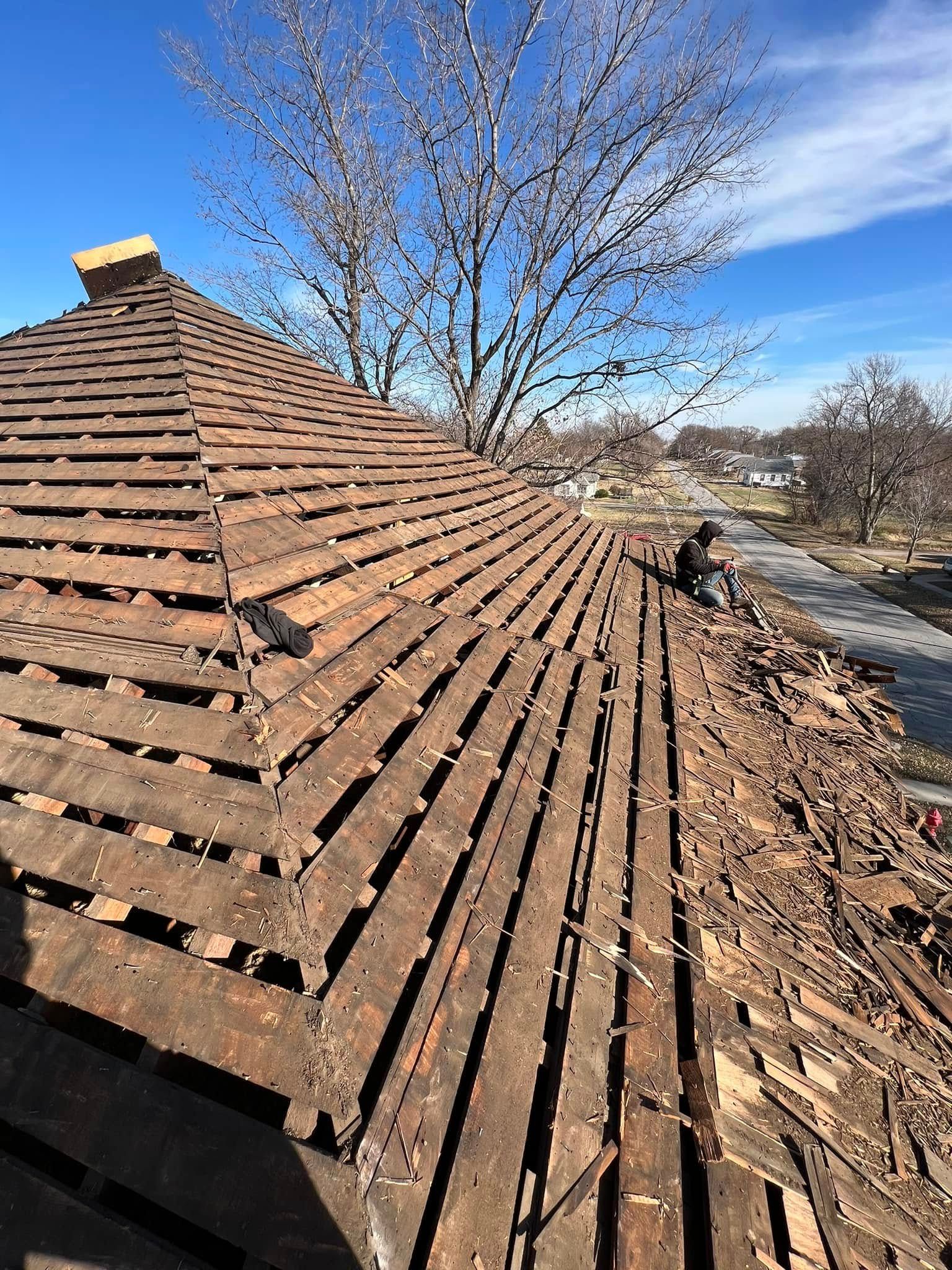 Damaged shingle roof with missing tiles and a tree in the background under a clear blue sky
