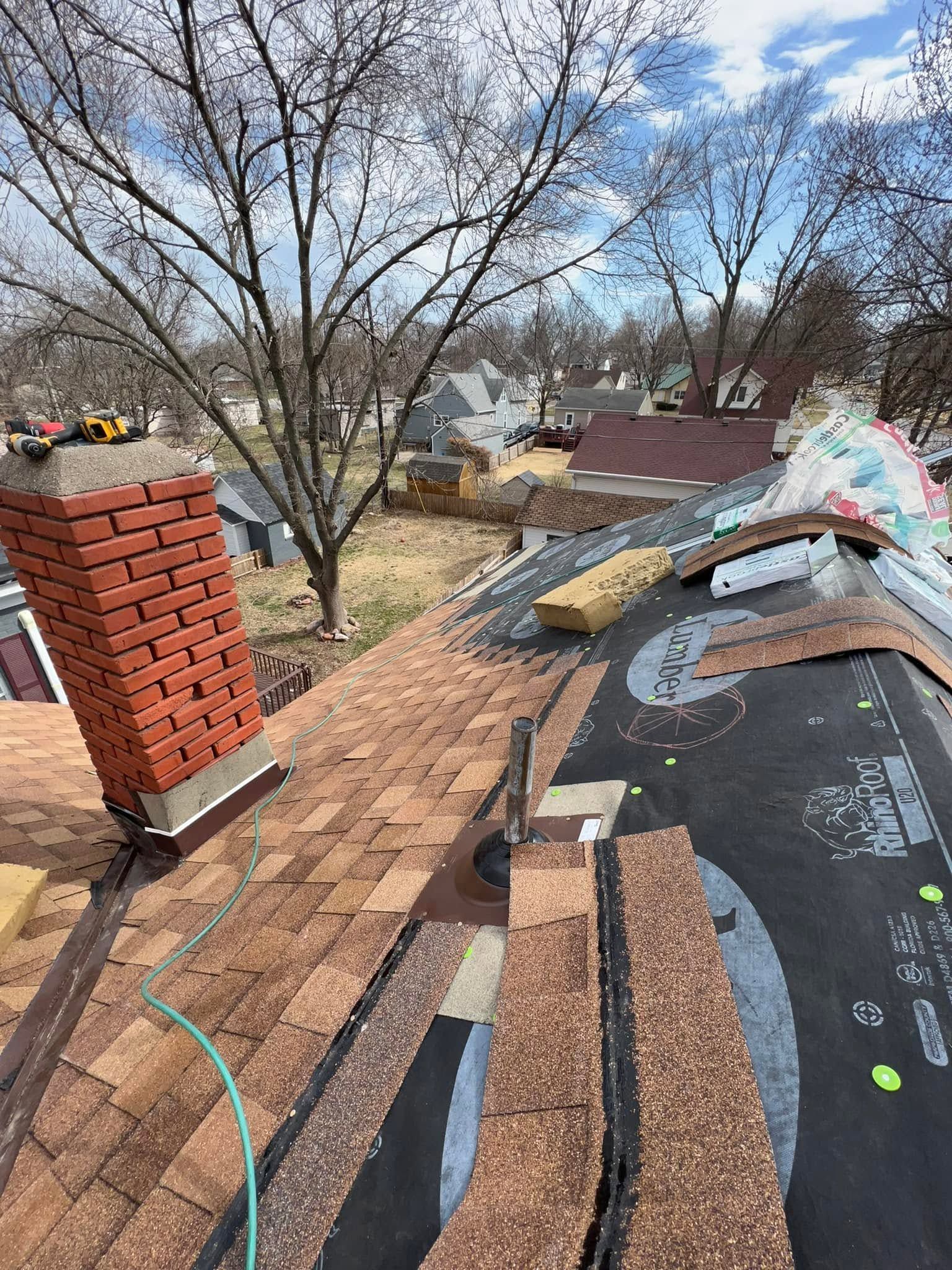 Roof repair with exposed shingles, brick chimney, and leafless trees under a blue sky.