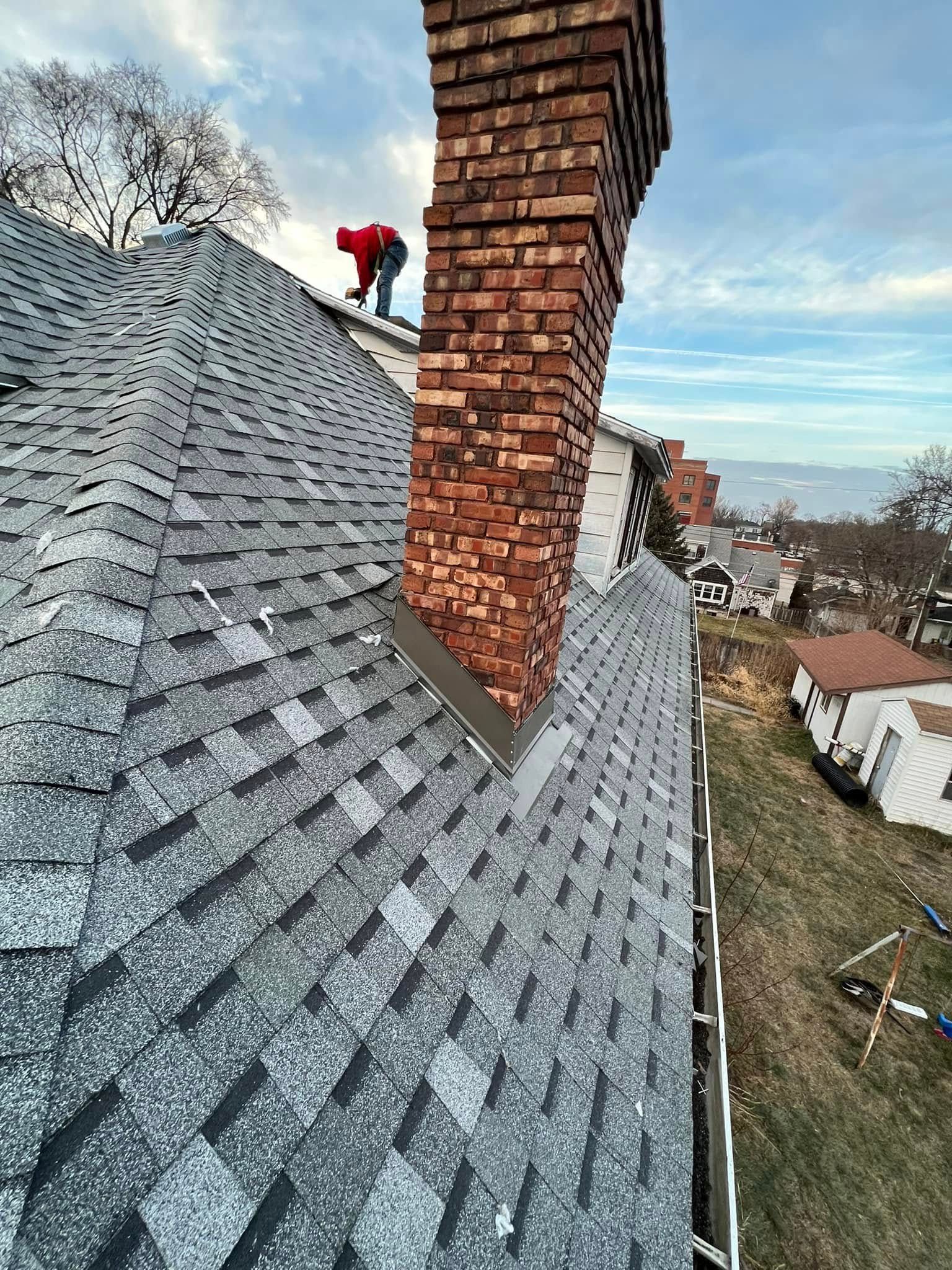 Worker inspecting gray shingle roof beside brick chimney on a sloped hillside homescape