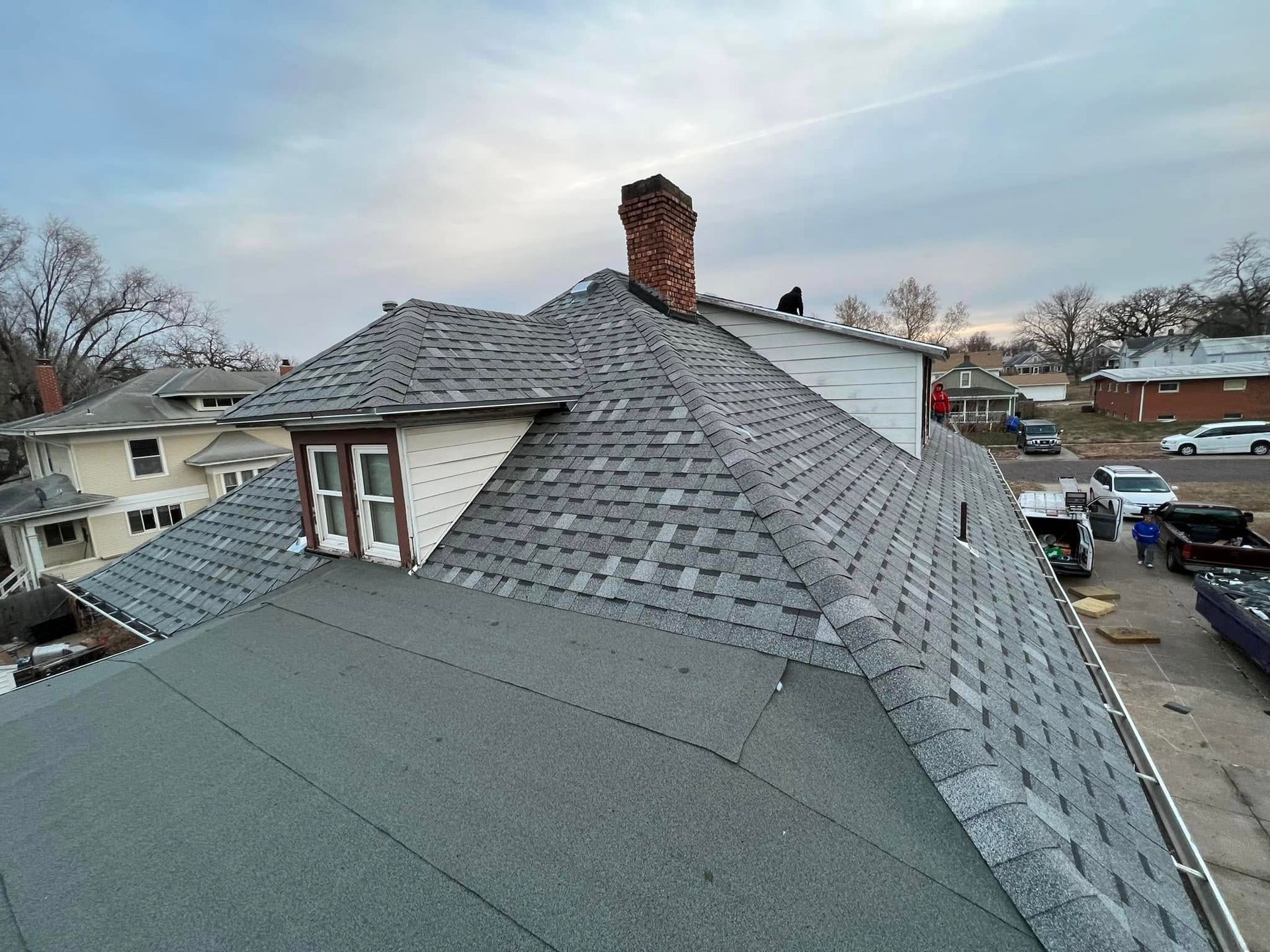 Rooftop with gray shingles and a brick chimney on a cloudy day, overlooking a residential neighborhood