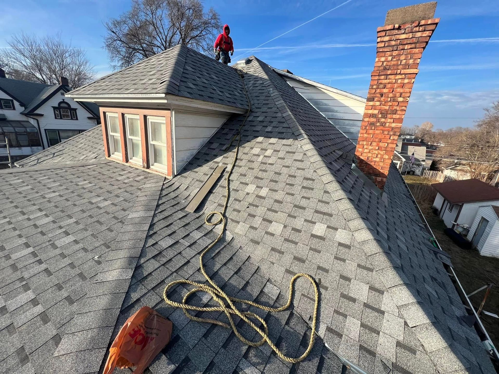 Worker on a gray shingled roof near a brick chimney, with tools and safety rope visible.
