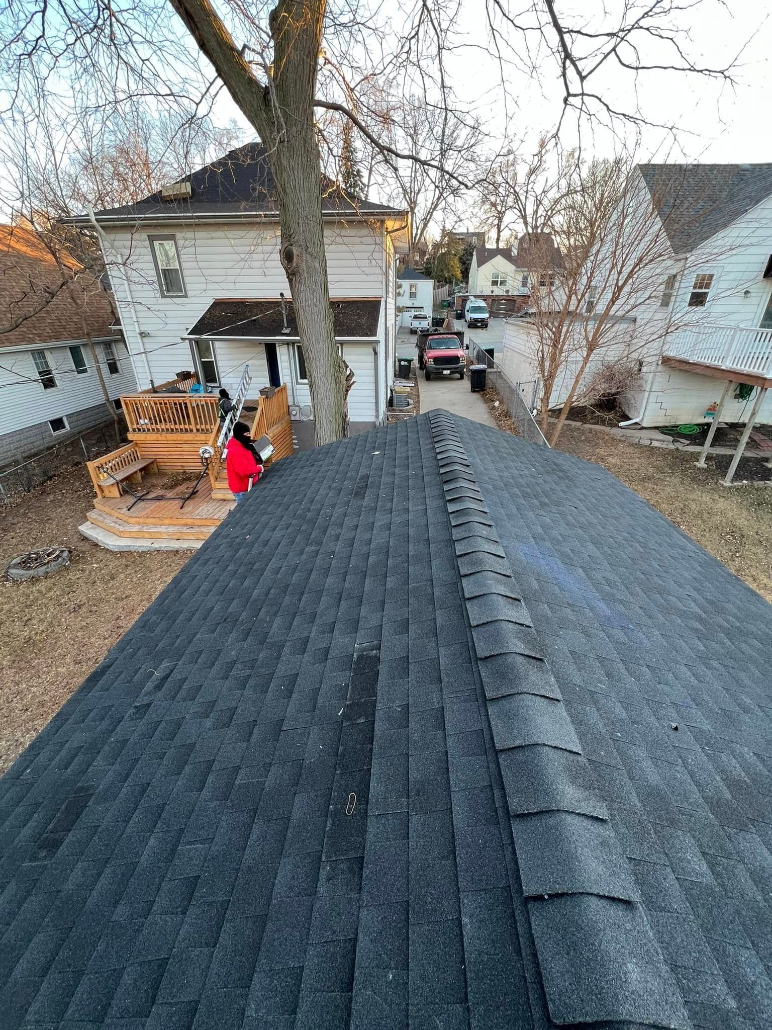 Rooftop view over dark shingle roof toward a backyard with houses, trees, and a person in red near a deck