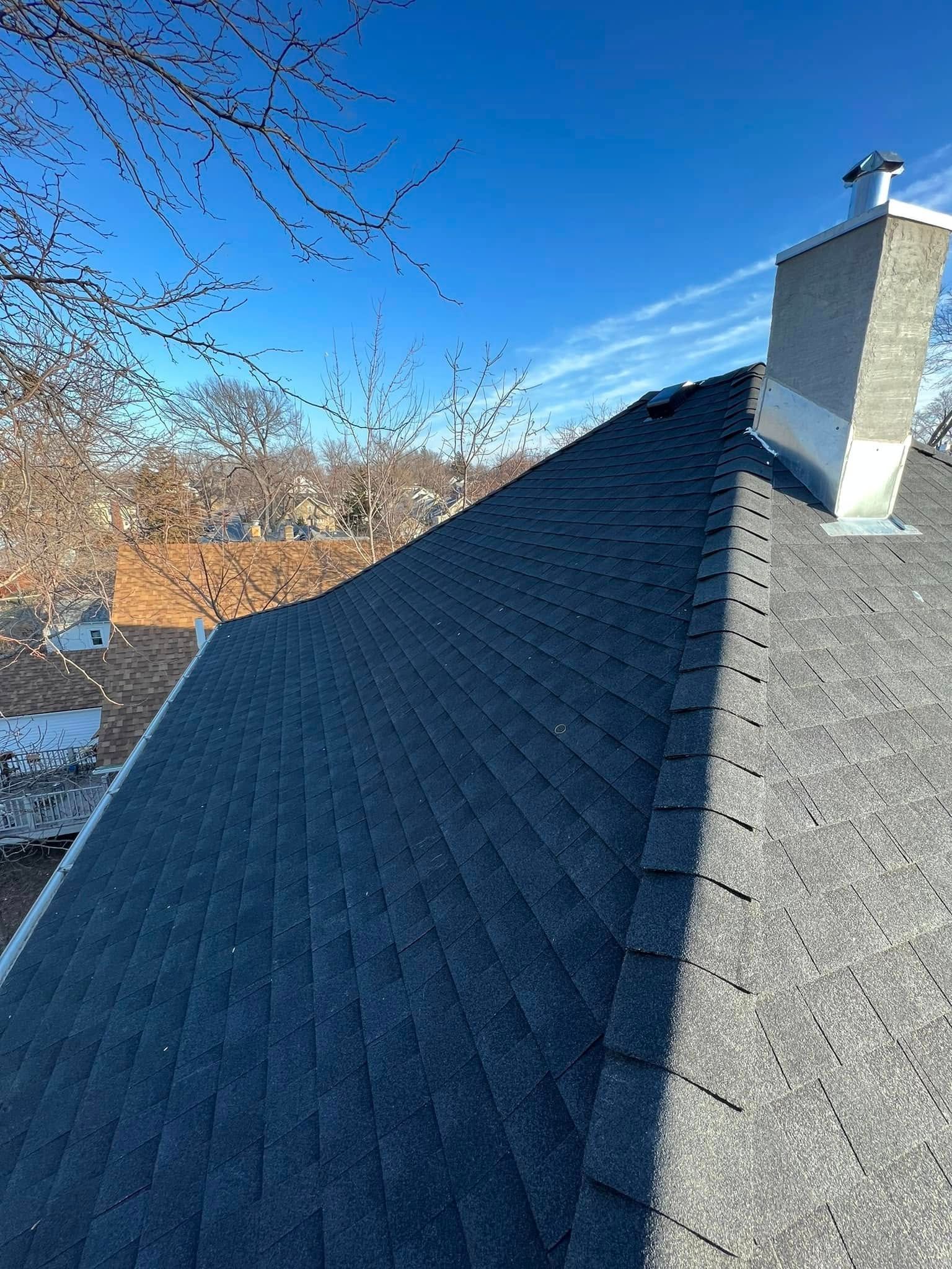 Blue-gray shingled roof with a chimney under a clear blue sky, with trees in the background