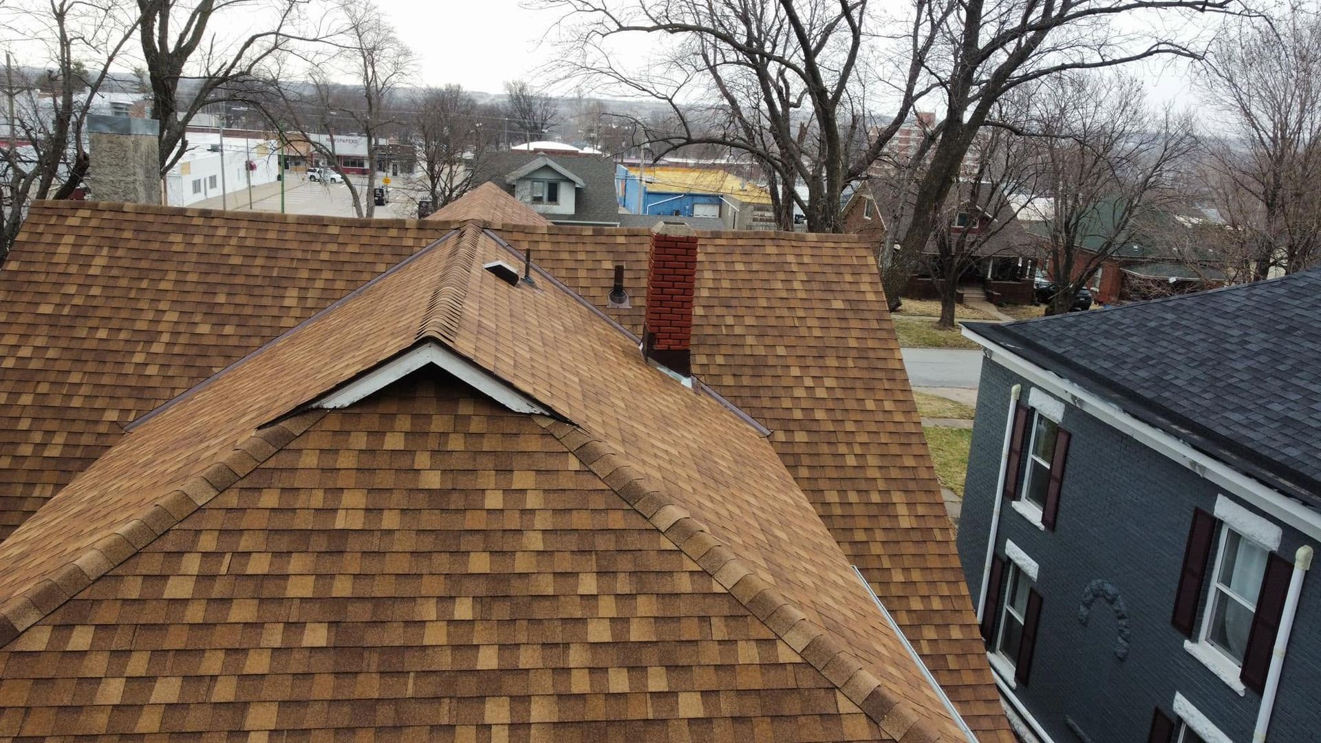 Rooftops with brown shingles beside a blue house, seen from above in a leafless neighborhood.