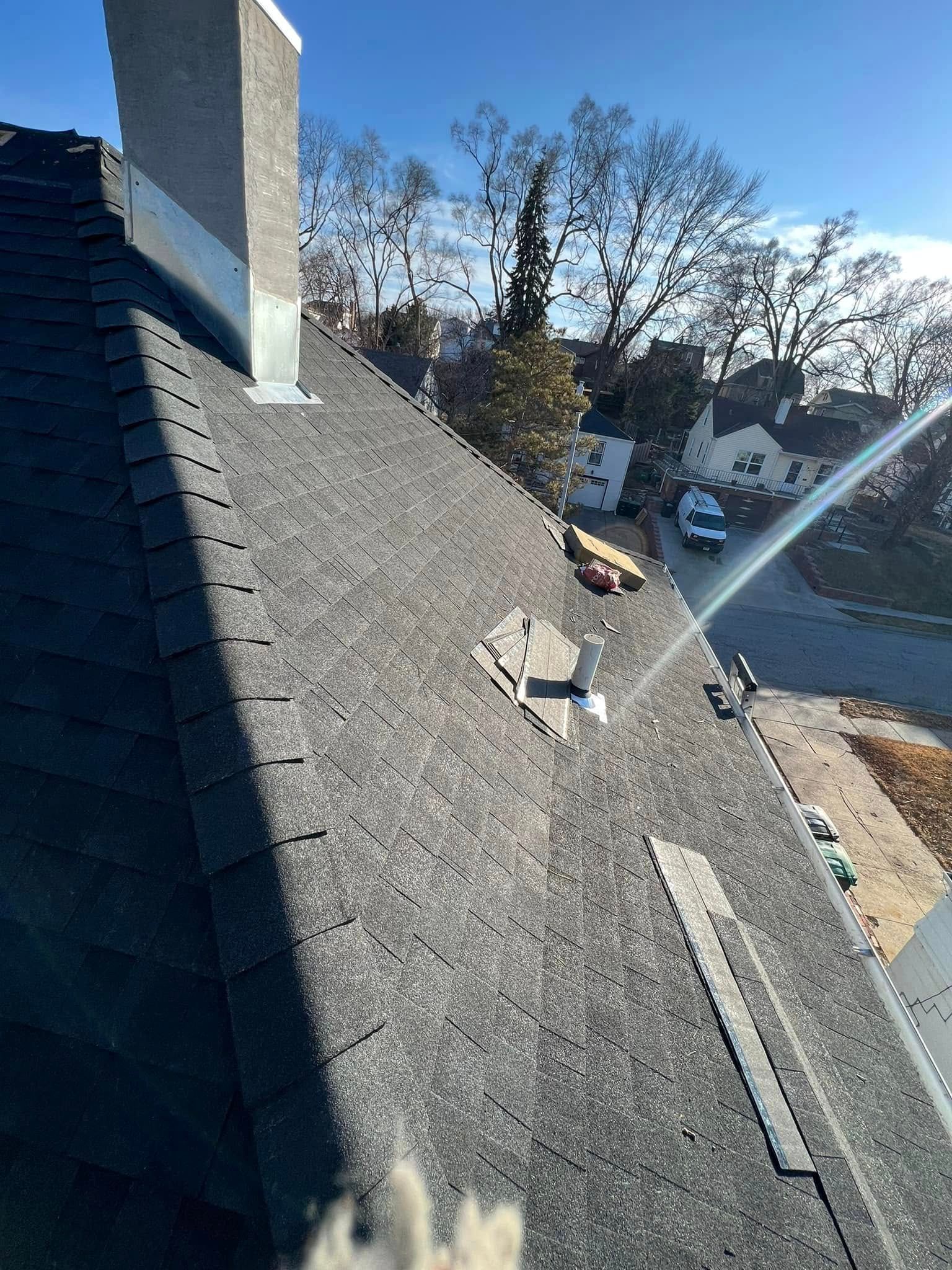 Roof with dark shingles, chimney, and vent pipe overlooking a street and trees under a bright sky