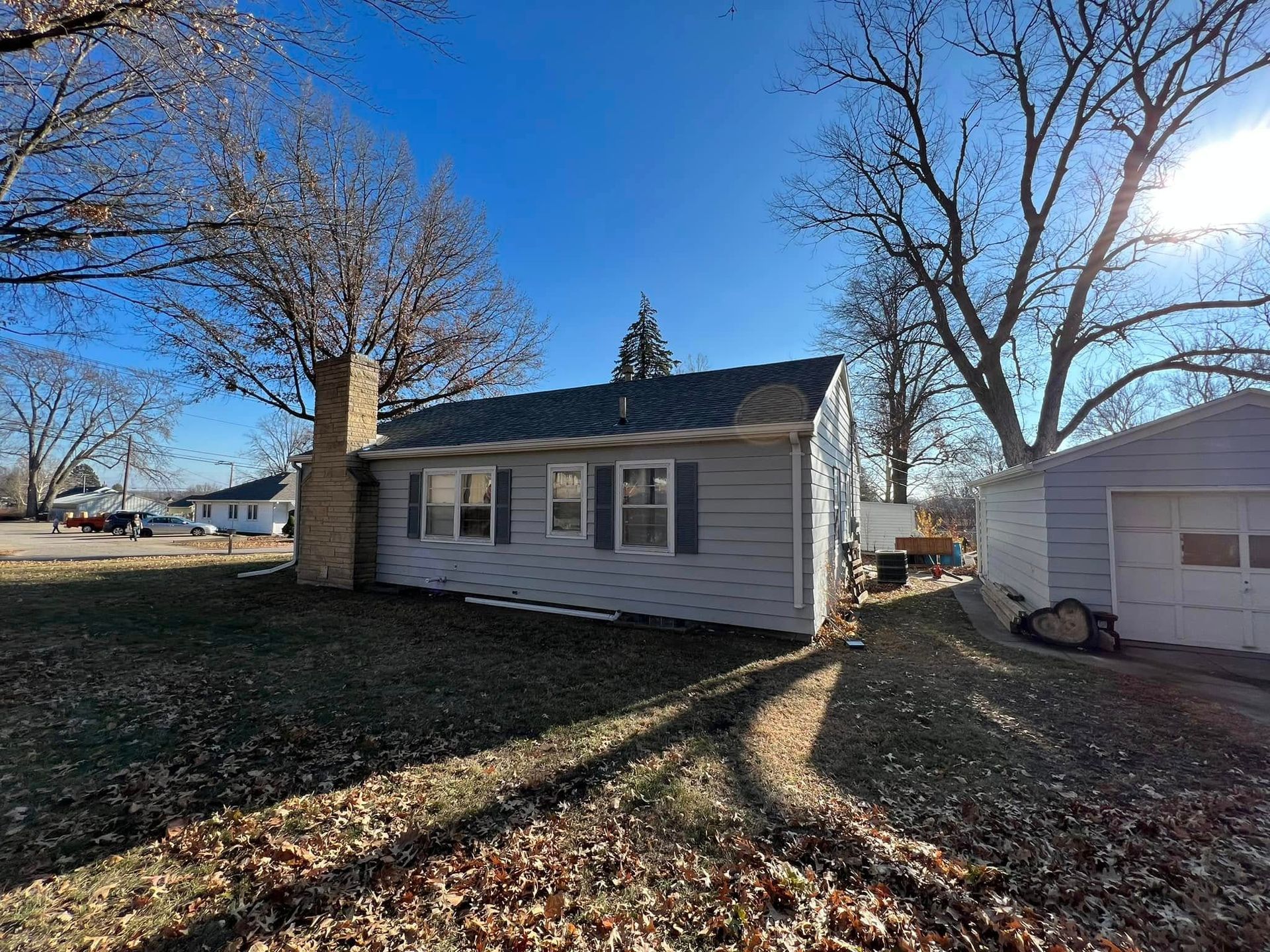 Small white house with a chimney, leafless trees, and a detached garage on a sunny day.
