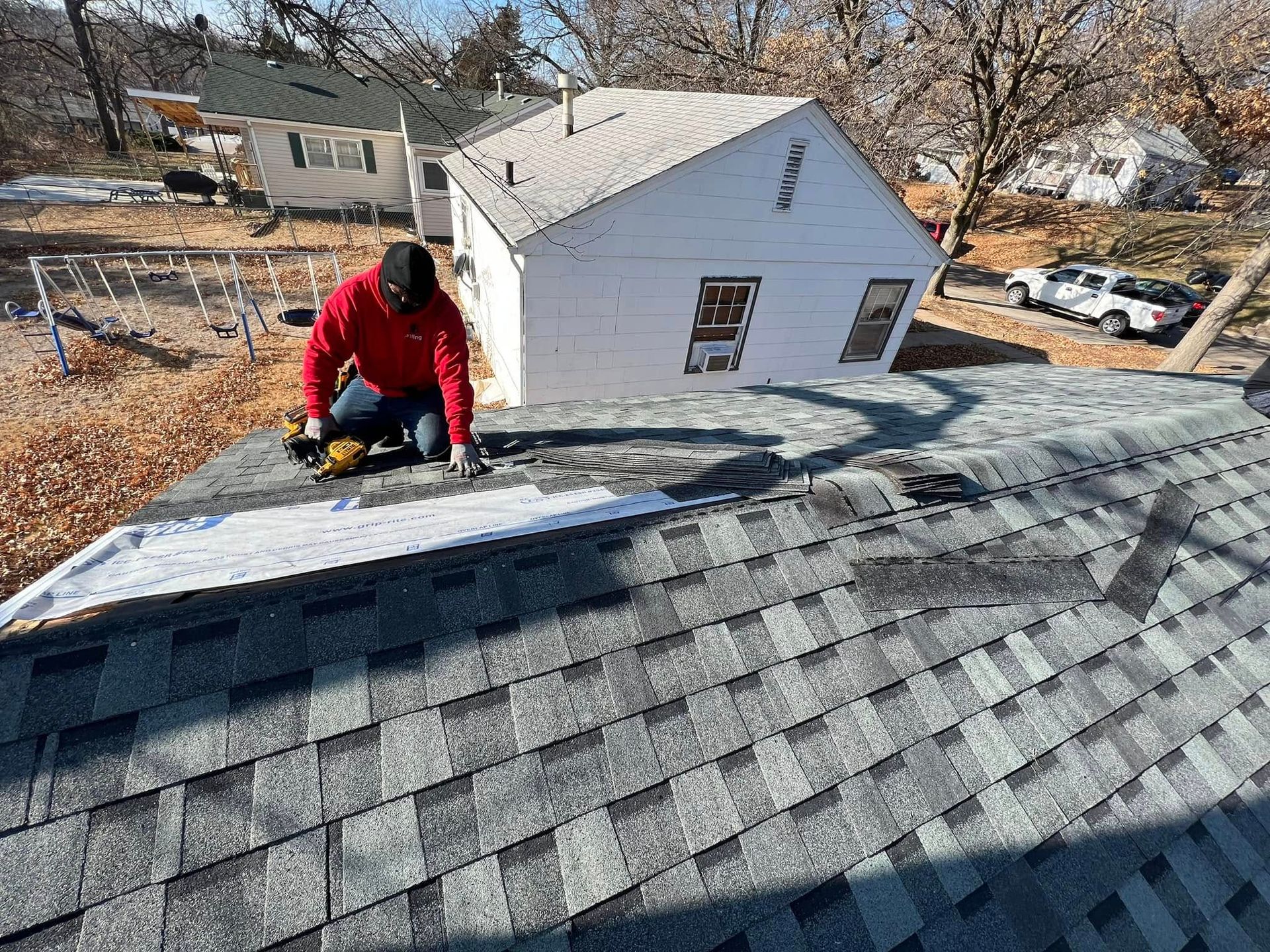 Worker in red jacket installing shingles on a gray roof beside a small white house.