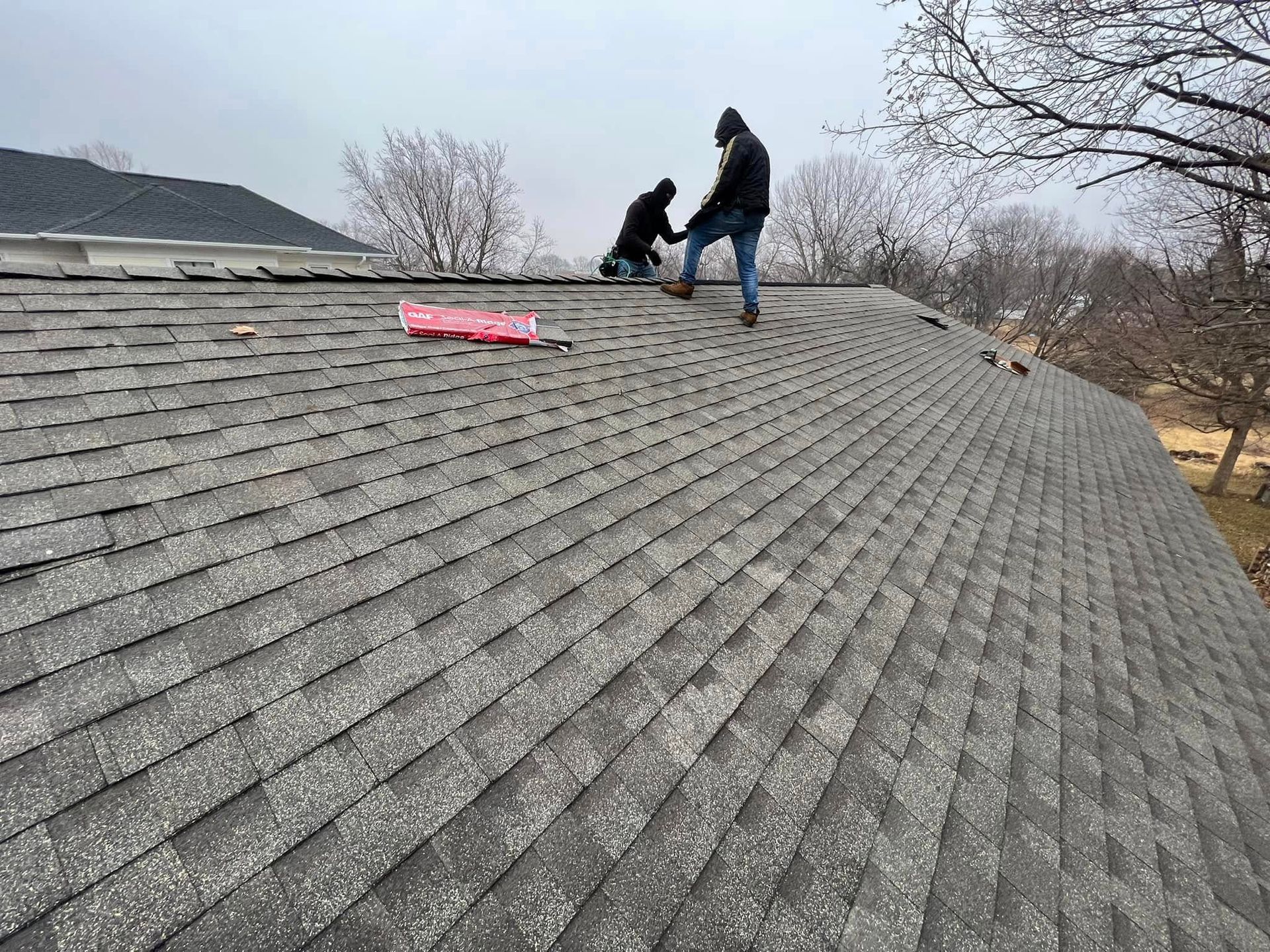 Two workers on a gray shingled roof, one holding a tool near a pink mat.
