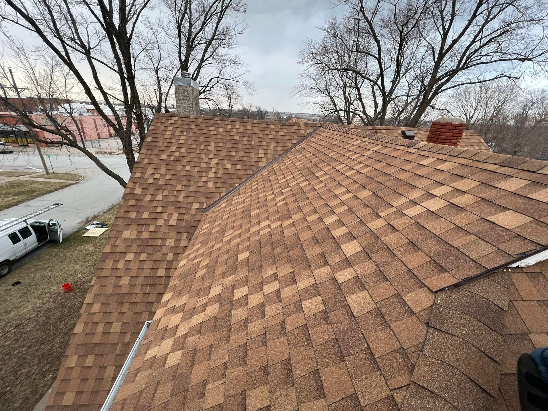Aerial view of a shingled roof with bare trees and a street nearby