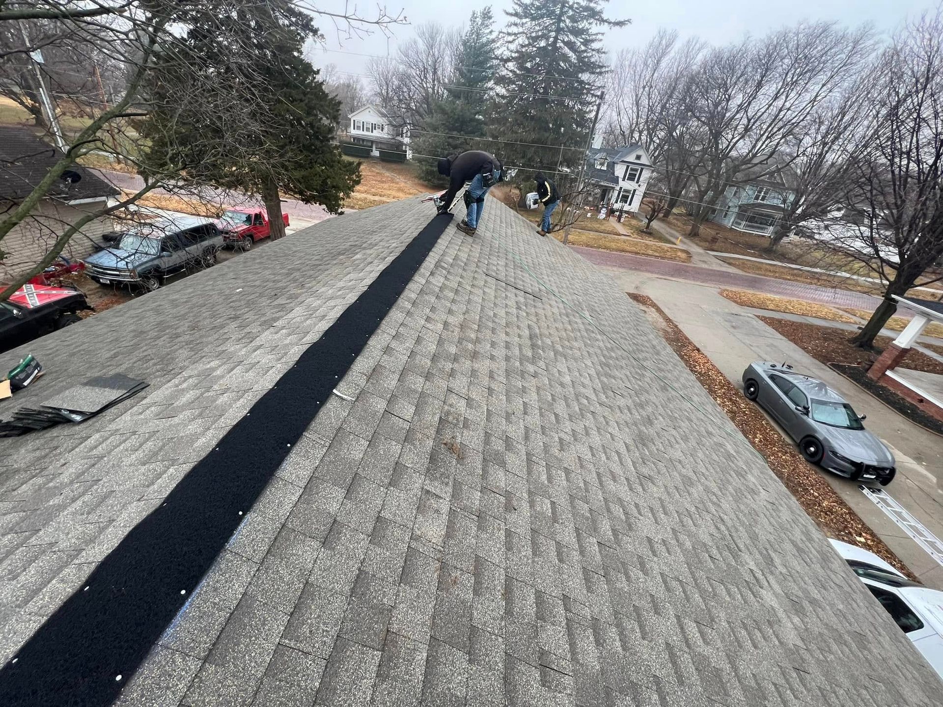 Worker on a rooftop with shingles and a long black strip, viewed toward a snowy residential area.
