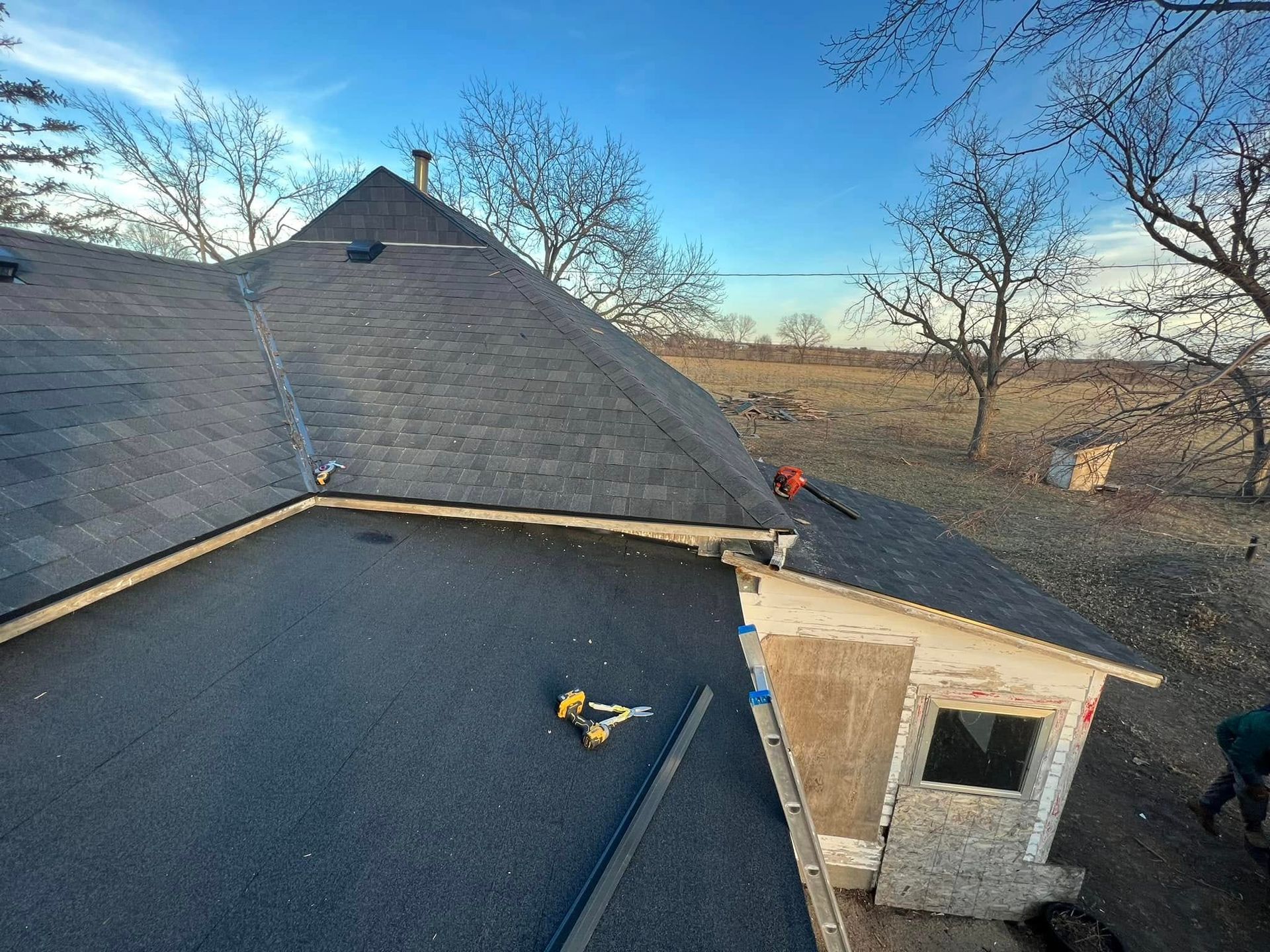 Roofer repairing a shingled roof beside a stone chimney in a rural yard, with bare trees and a blue sky