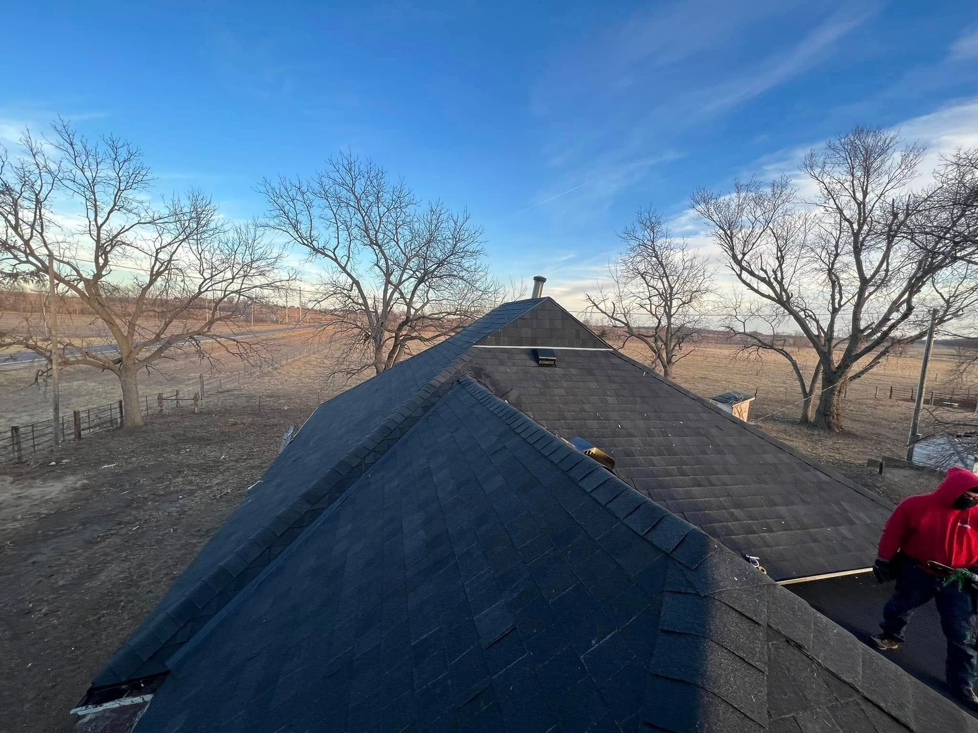 Roof with black shingles in a rural landscape, with a person in a red jacket working on the right side.