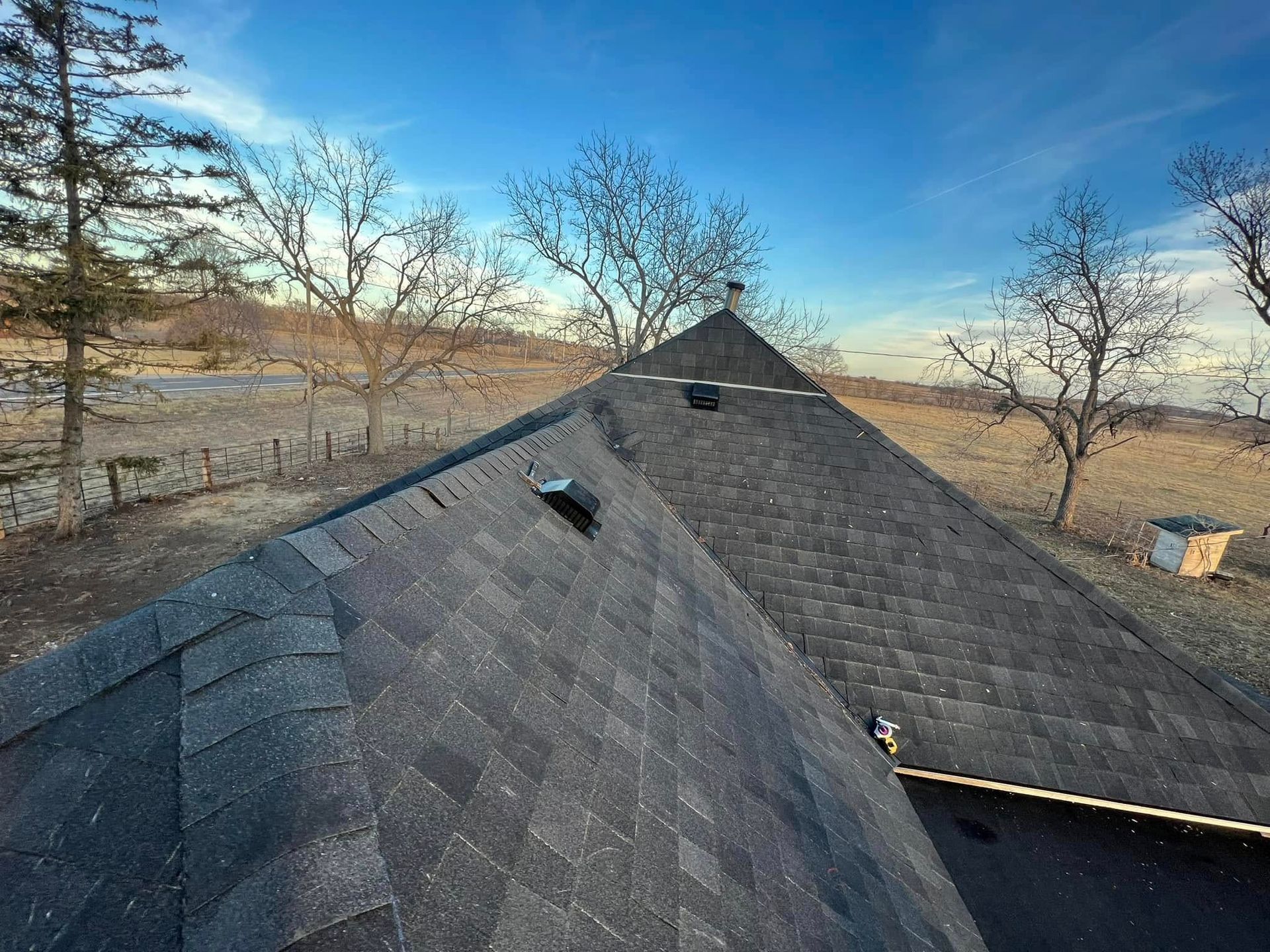 Dark gray shingled roof ridge under a blue sky, with leafless trees and fields in the background