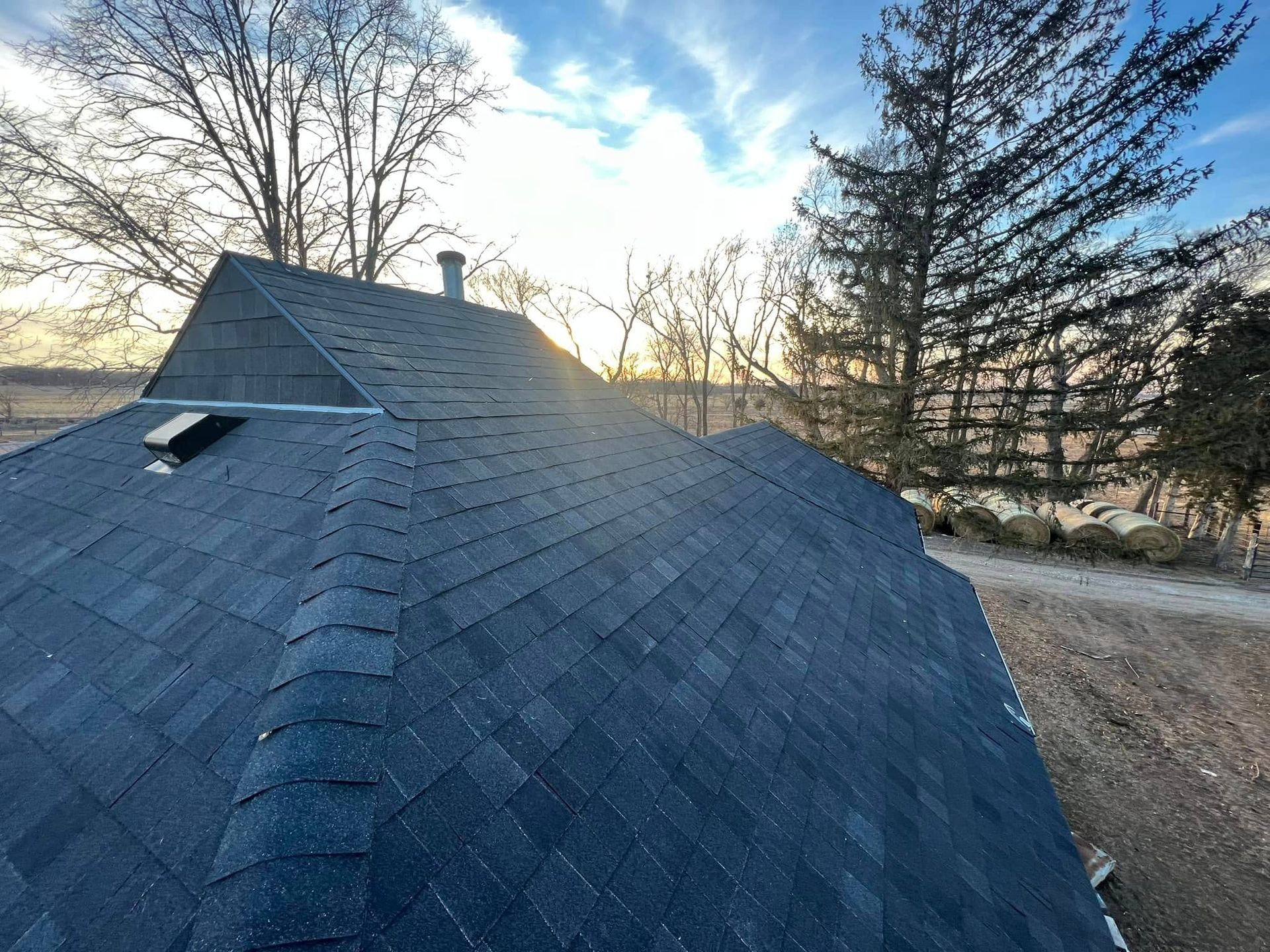 Roof shingles on a house at sunset, with trees and a gravel driveway in the background