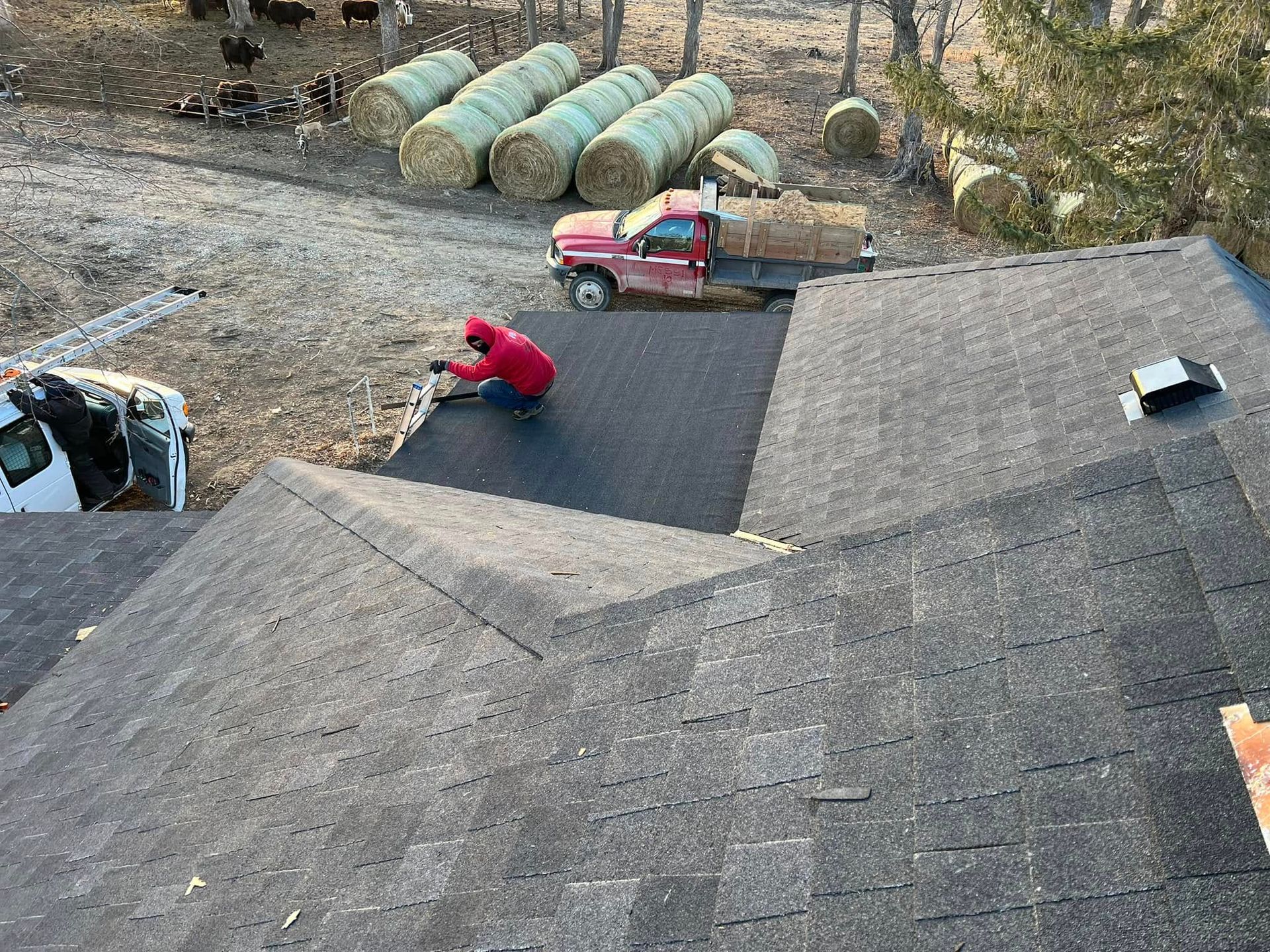 Worker in red shirt installing shingles on a roof, with truck and hay bales in the background.