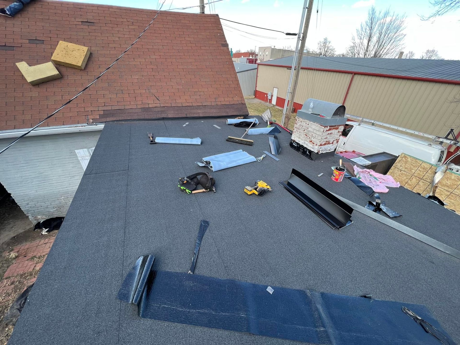 Flat rooftop under repair with roofing tools, shingles, and materials scattered across dark underlayment.