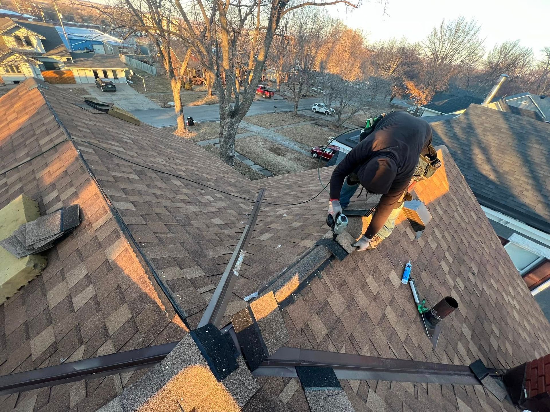 Worker on a shingled roof using a power tool near a ridge vent, with ladders and trees in the background
