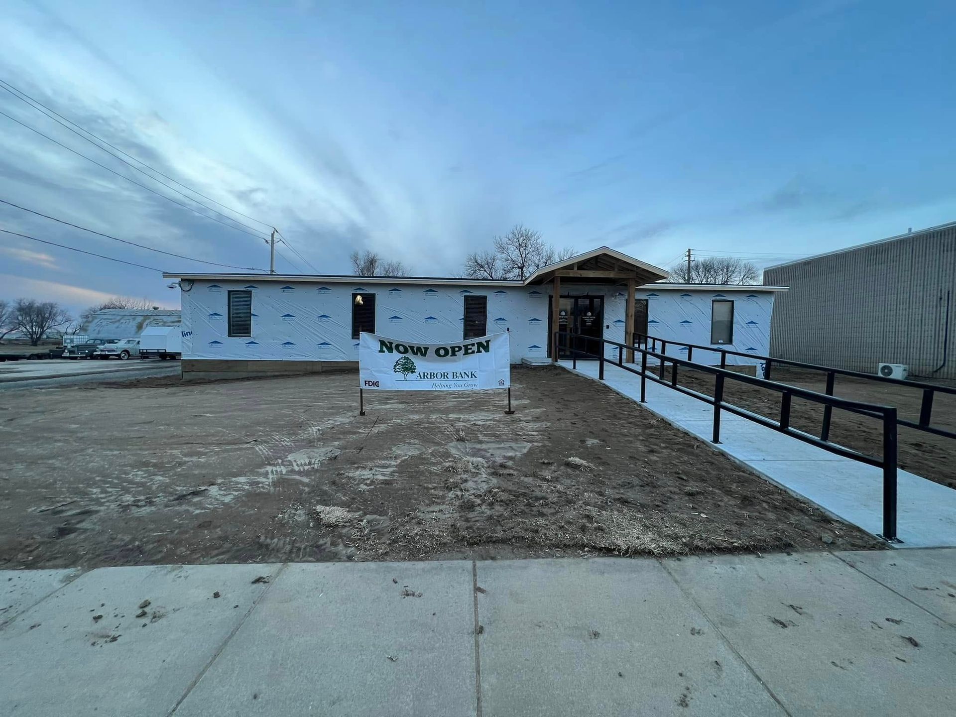 Single-story white house with a dirt yard, concrete sidewalk, and ramped walkway under a cloudy blue sky