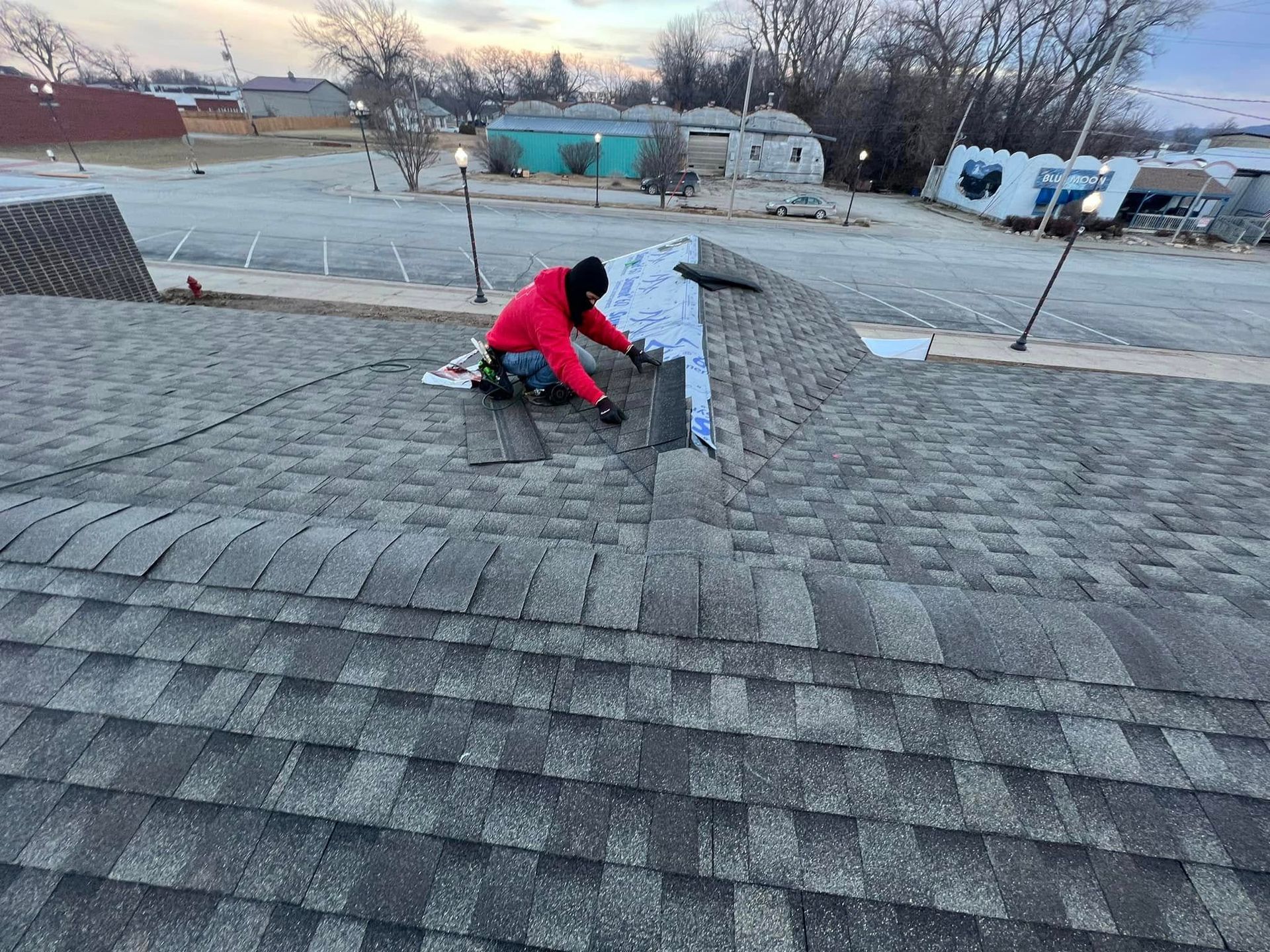 Worker in red jacket repairing a shingled rooftop near a vent pipe at dusk
