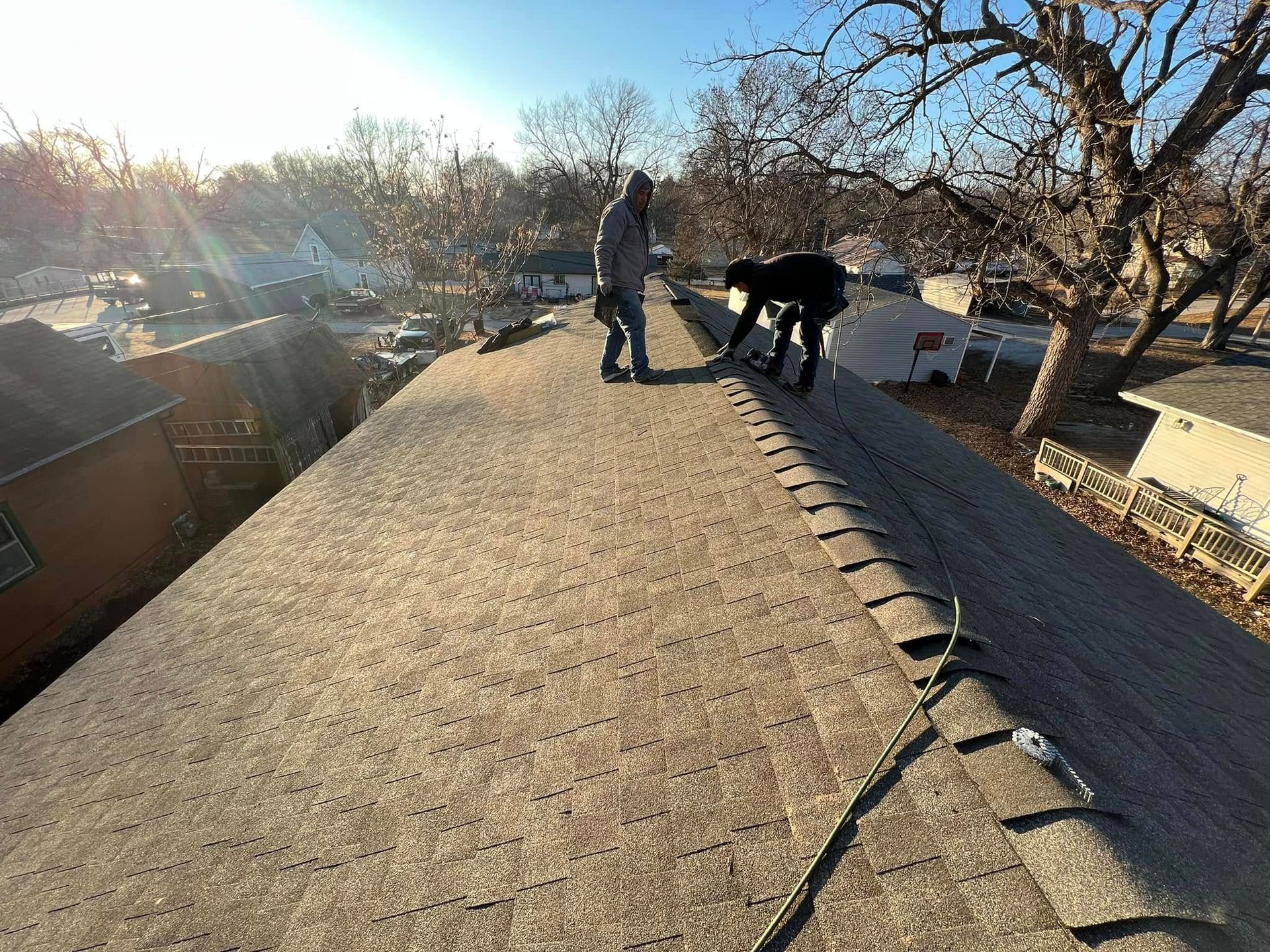 Two people on a rooftop, one bending near the edge in bright sunlight with trees and houses below.