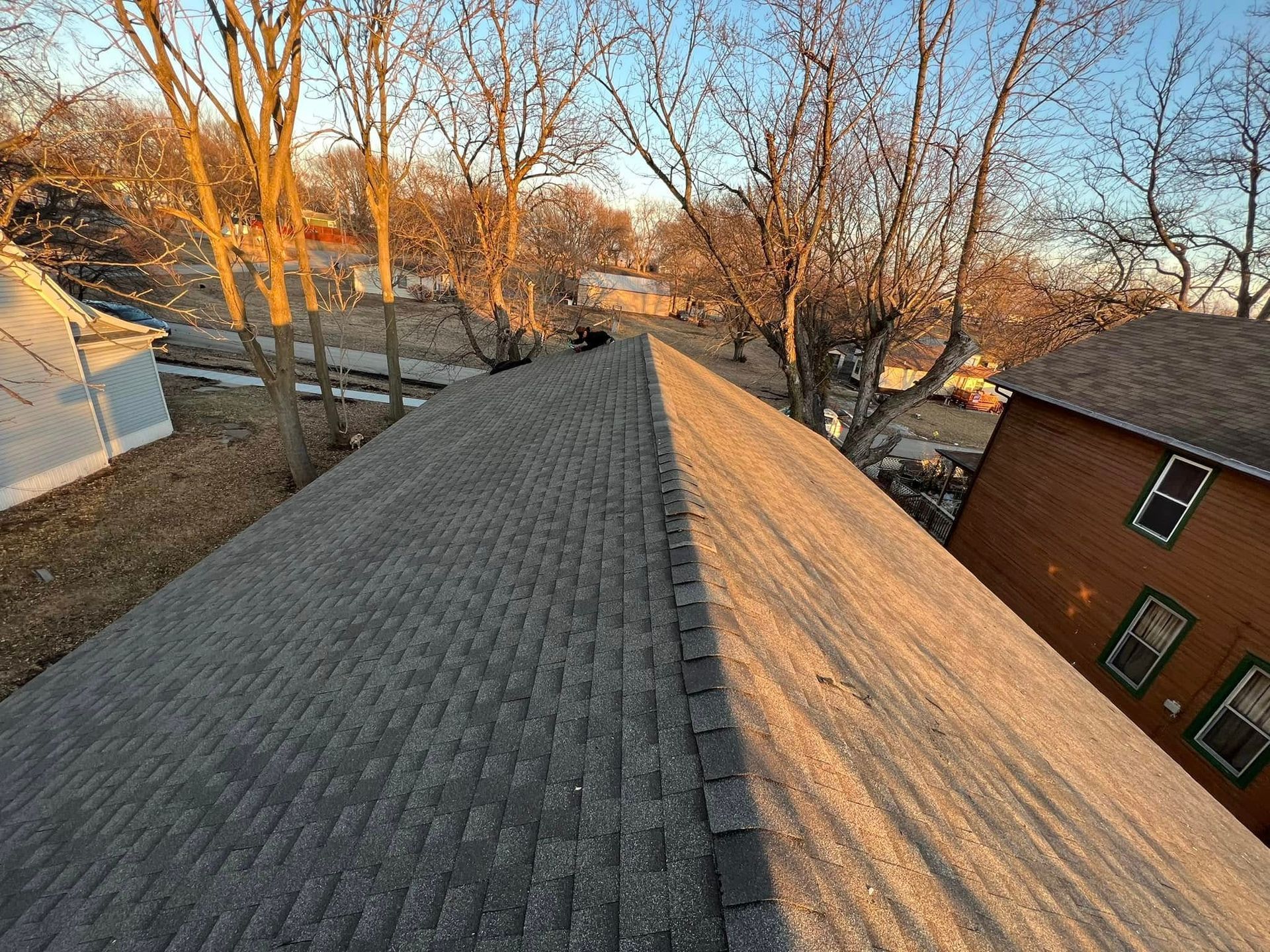View from a rooftop ridge at sunset, showing gray shingles, bare trees, and nearby houses.
