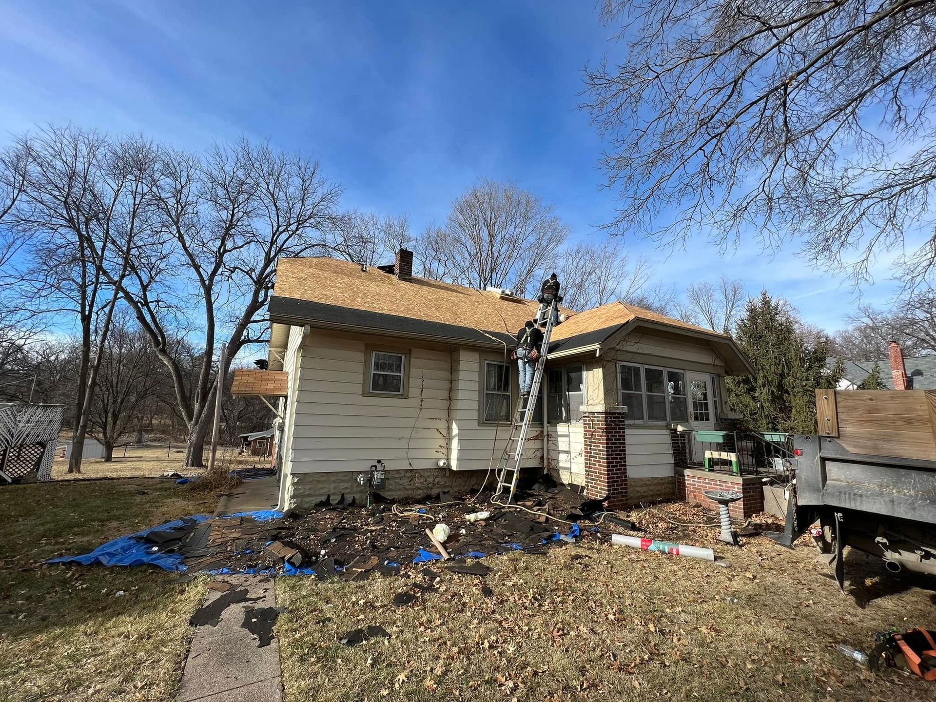 Small beige house with damaged roof, muddy yard, and scattered debris under a blue sky.