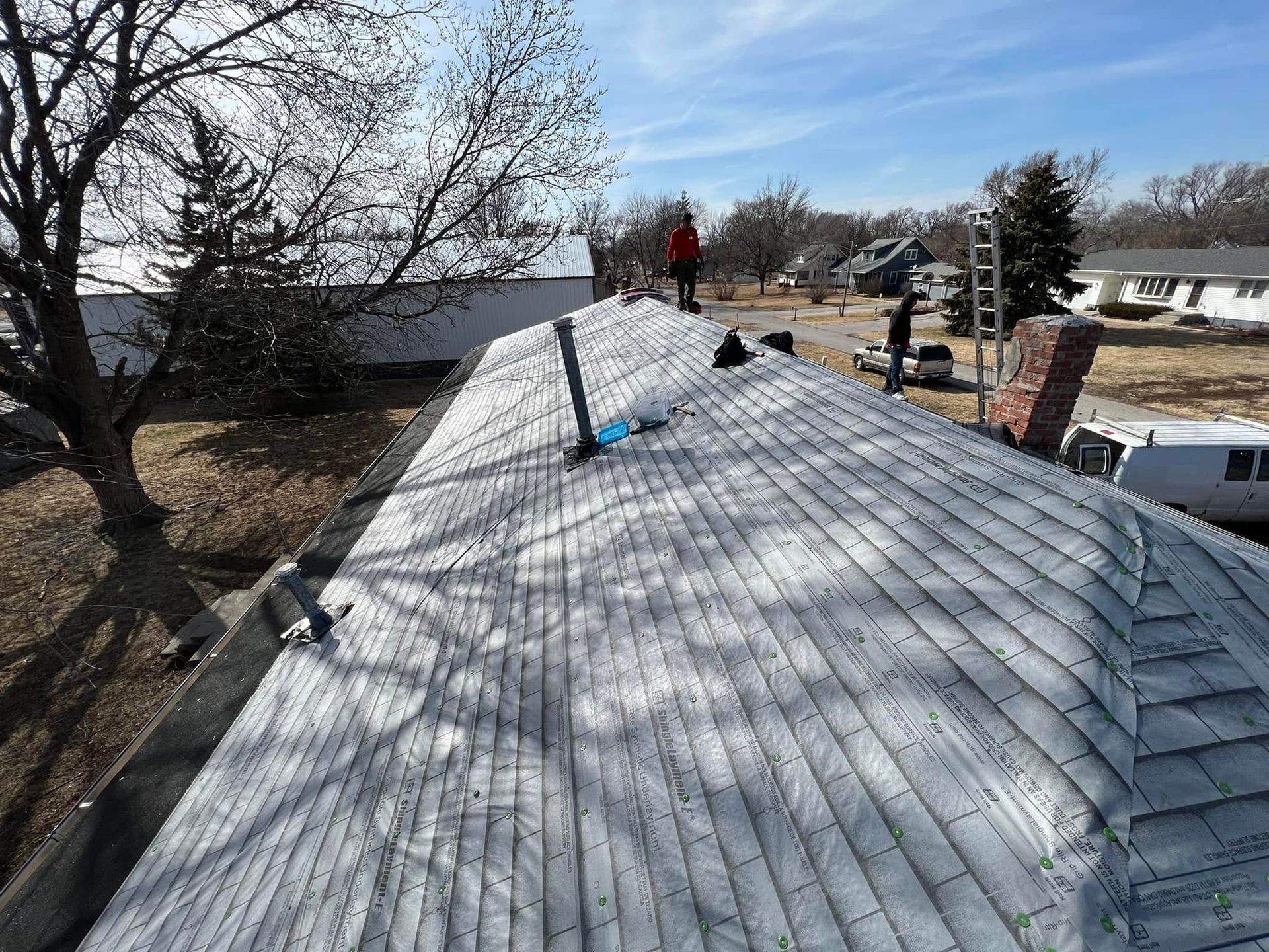 Workers repairing a gray shingle roof on a house, with ladders and trees nearby