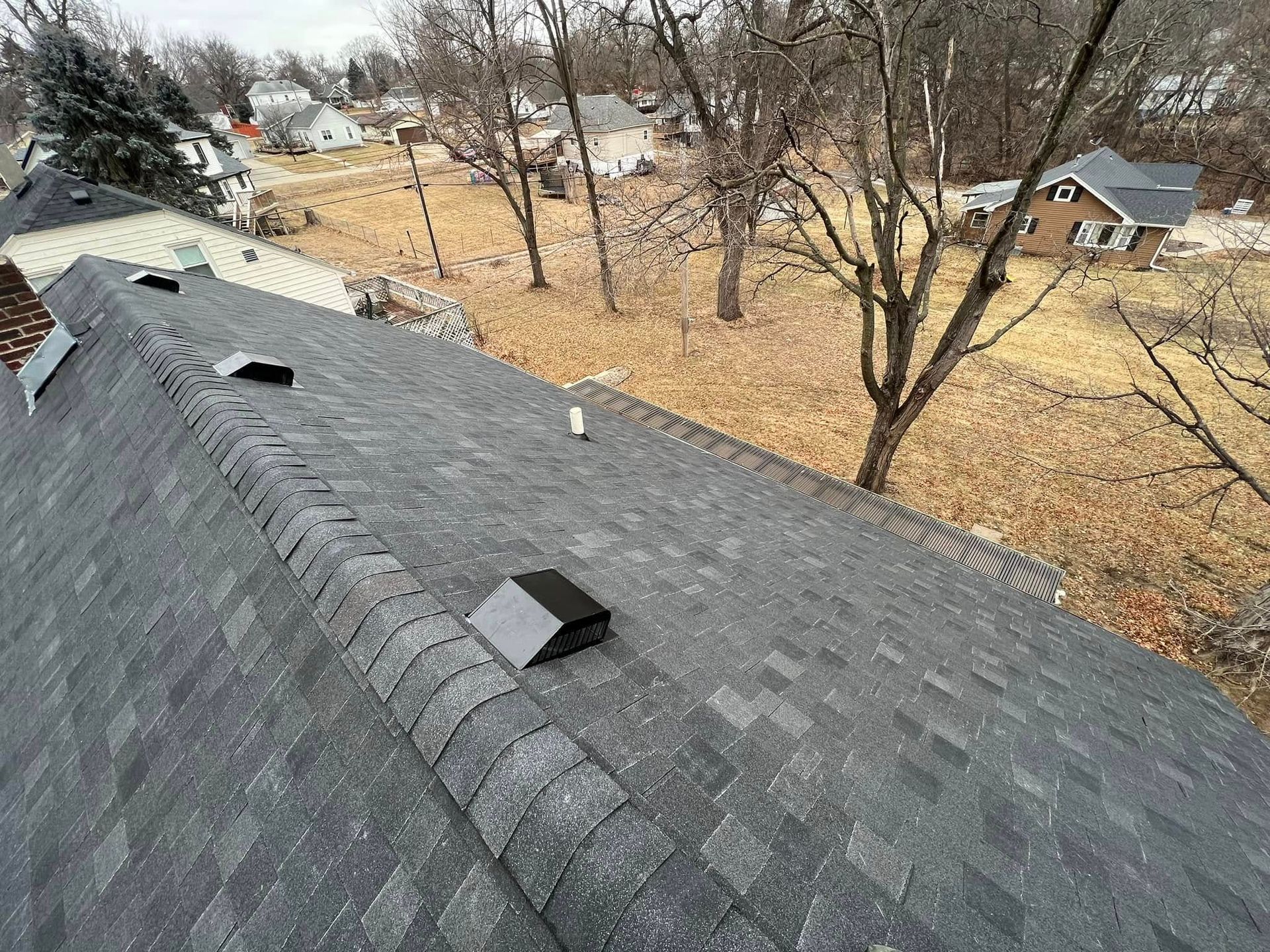 Shingled roof with two vents overlooking a leaf-covered yard and trees