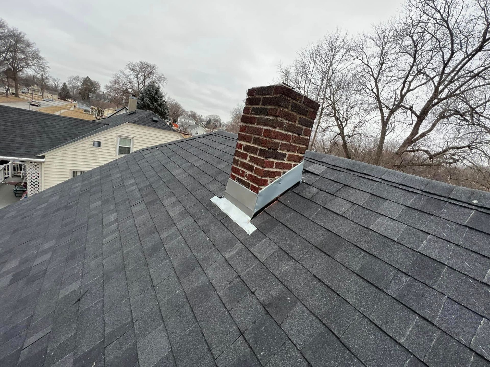 Rooftop with gray shingles and a brick chimney flashing repair, with houses and leafless trees in the background