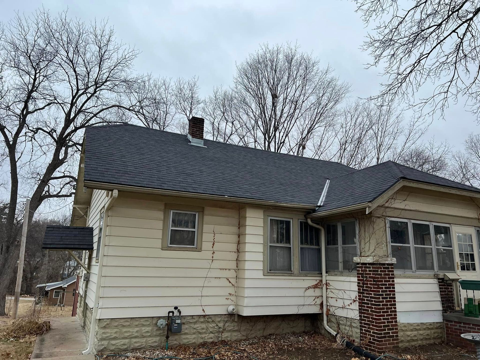 Small beige house with a dark roof and brick porch in a leafless yard under cloudy sky
