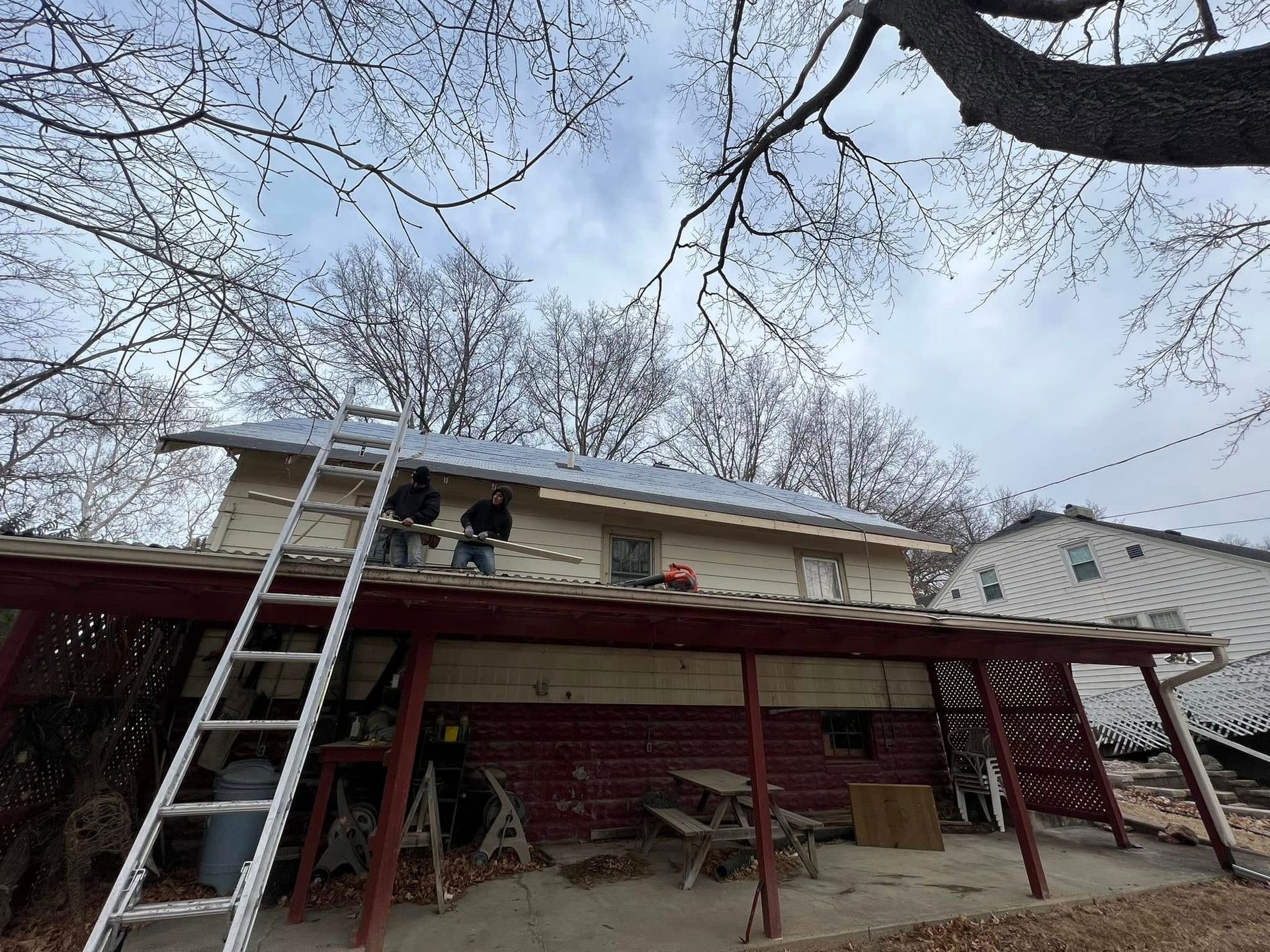 Ladder leaning against house roof under repair, with red deck and bare trees overhead