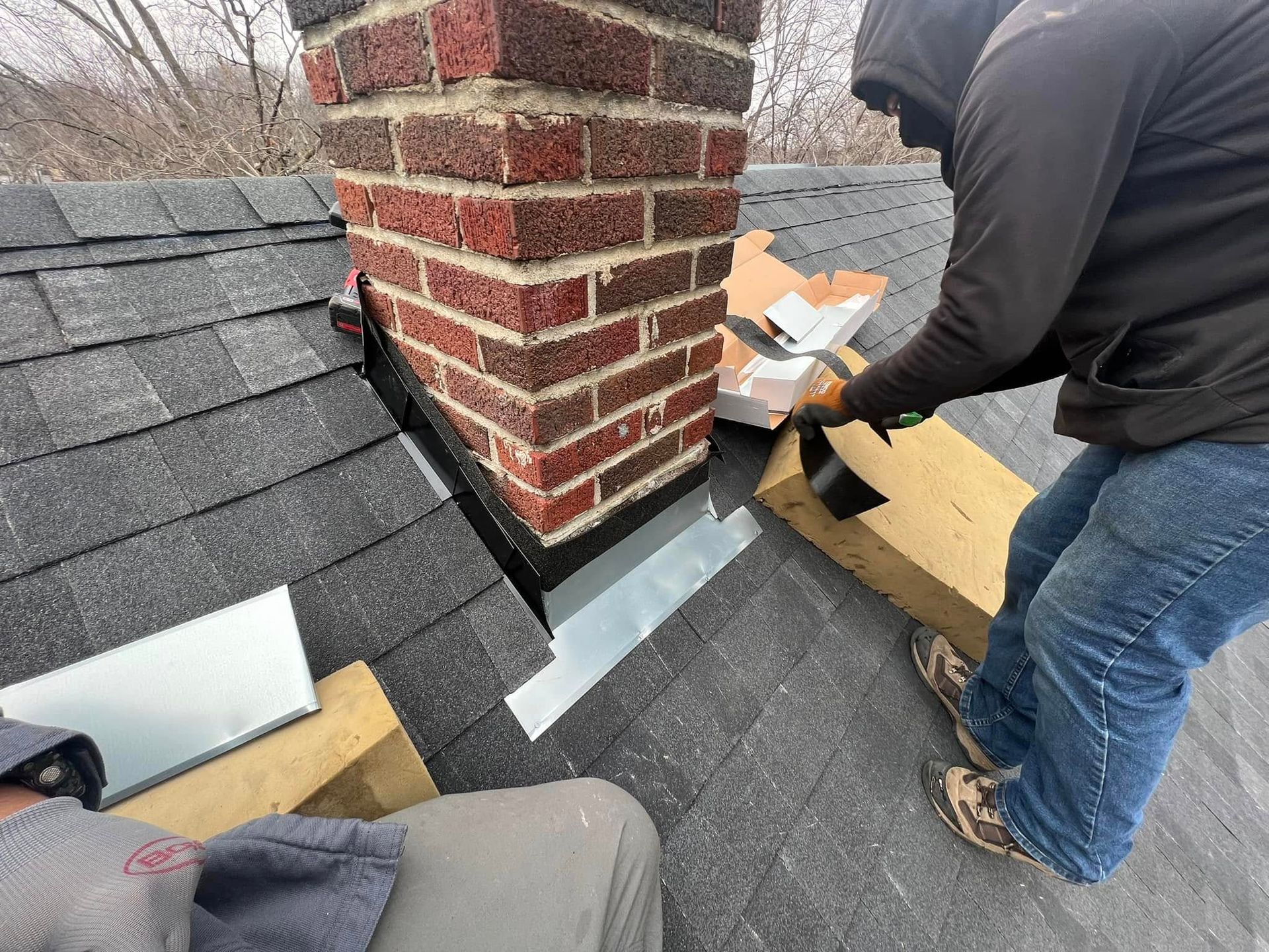 Two workers installing roof flashing around a brick chimney on a shingled roof