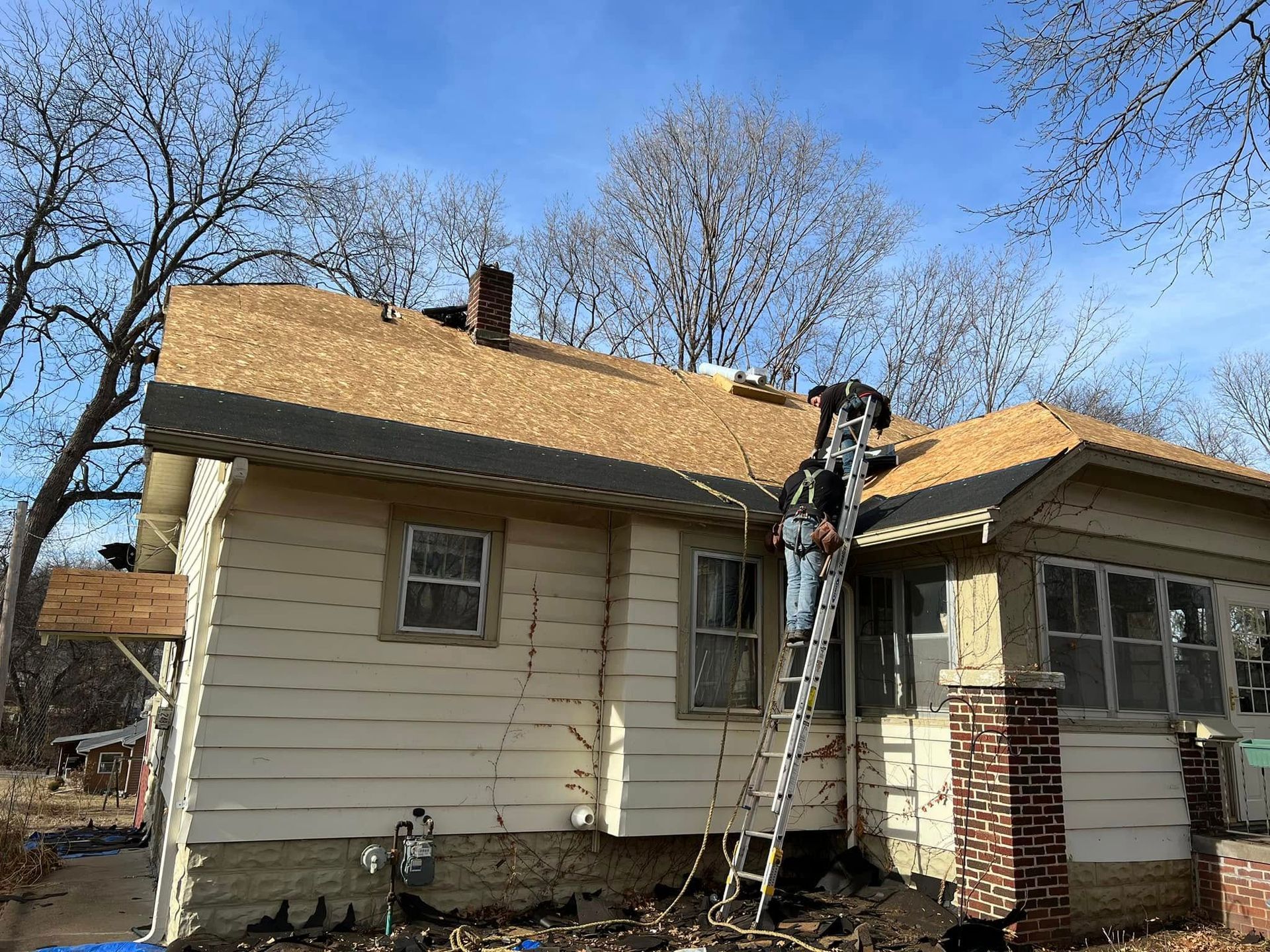 Worker on a ladder repairing the roof of a beige house with a brick chimney.