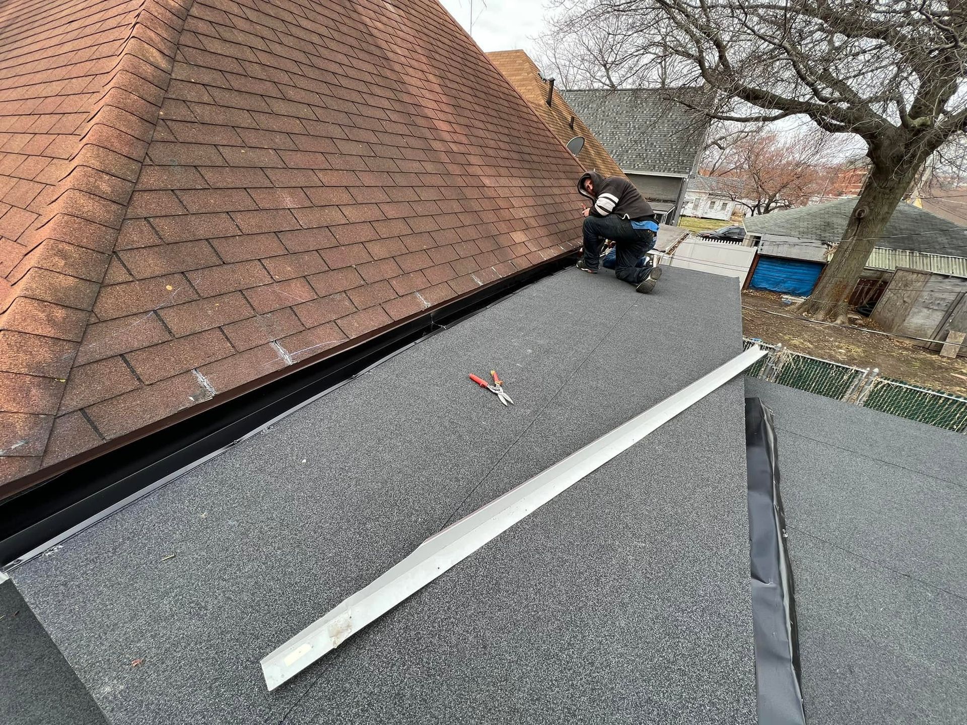 Worker on a flat roof beside a brick wall, installing roofing material with tools nearby.
