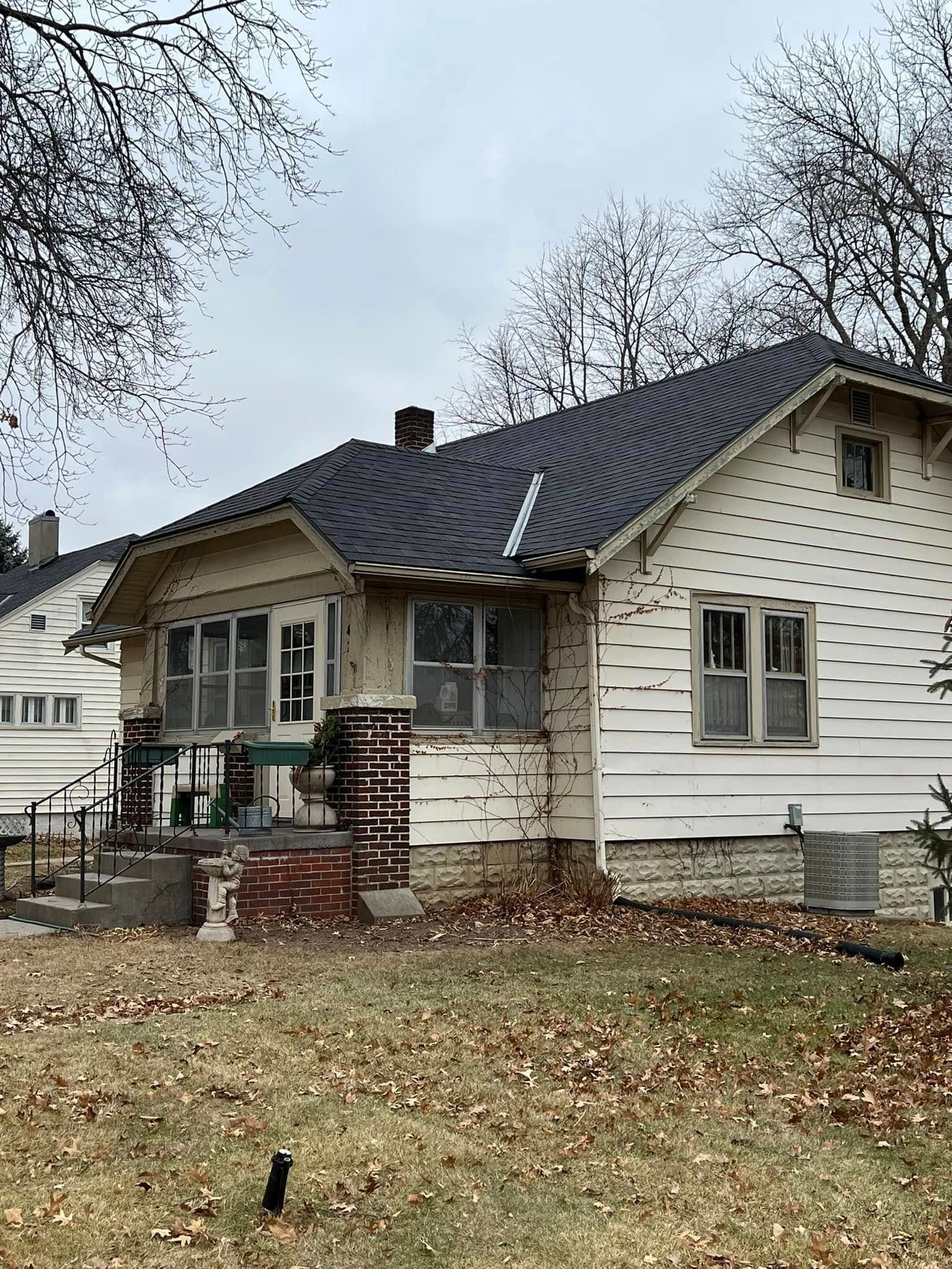 Small white house with dark roof and porch, front yard covered in leaves, seen on an overcast day